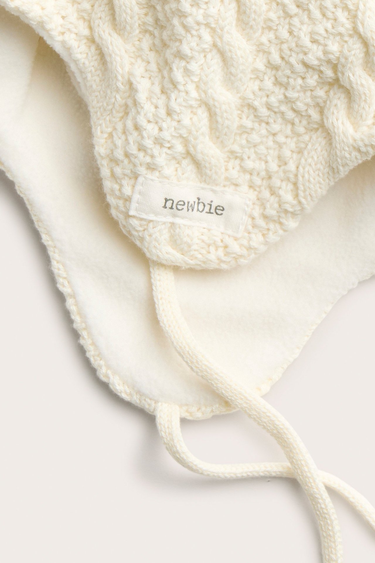 Nahaufnahme einer weißen Zopfstrick-Babymütze mit Fleecefutter und Bindebändern.