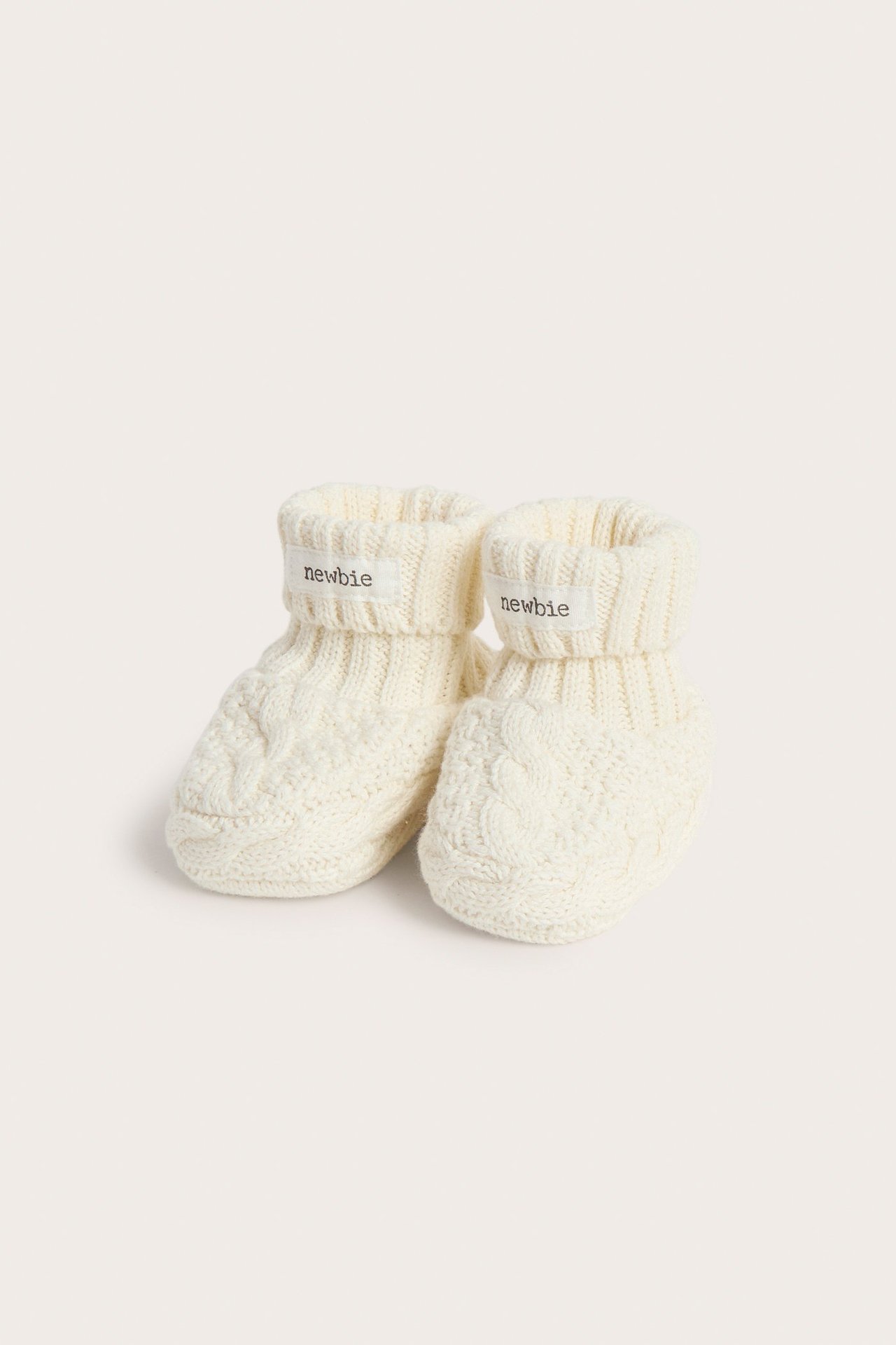 Front view of white cable-knit baby booties with ribbed cuffs and "newbie" labels.