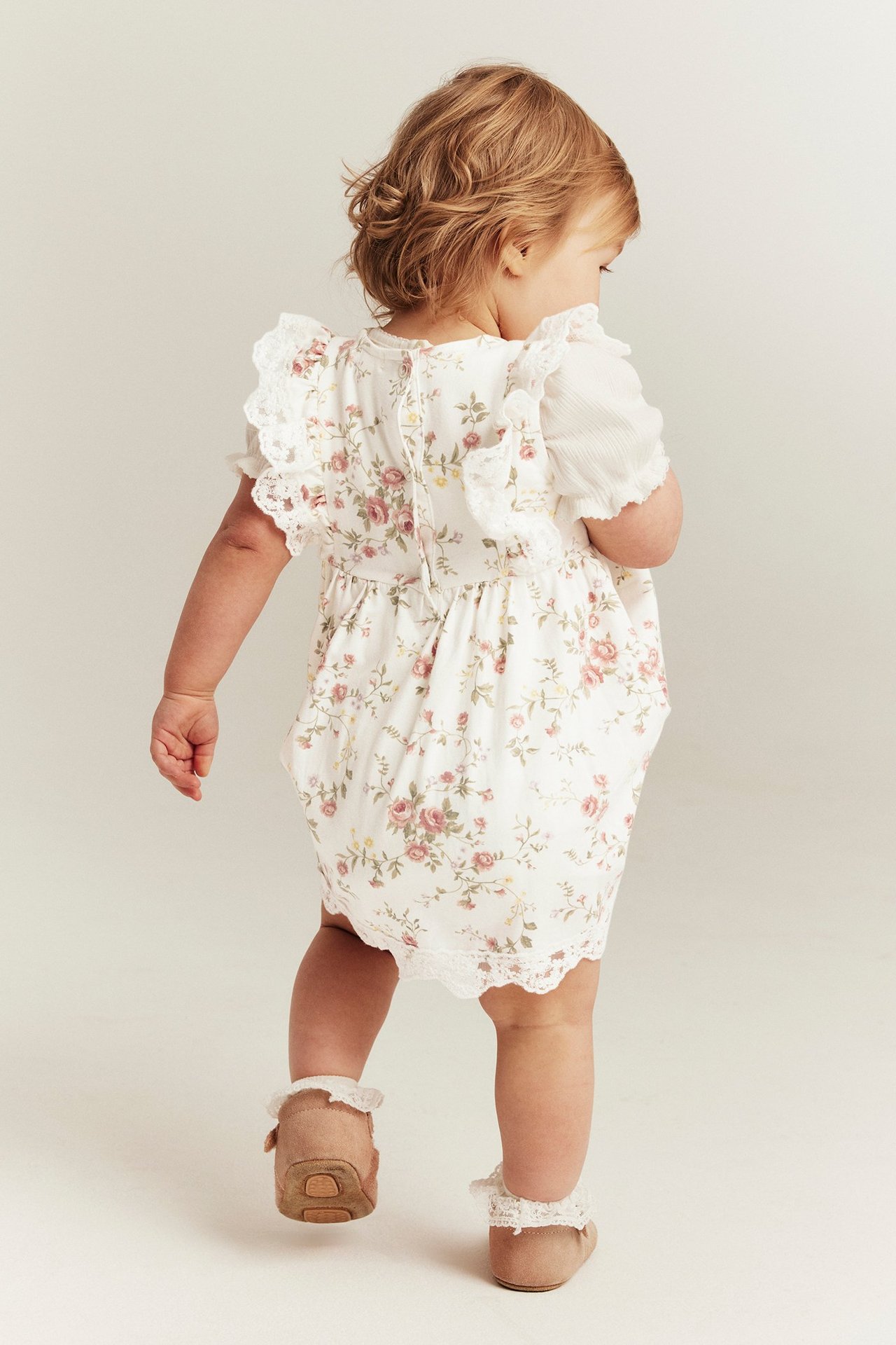 Rear view: Baby wears off-white floral sleeveless dress with lace trim, layered over white top, brown shoes.