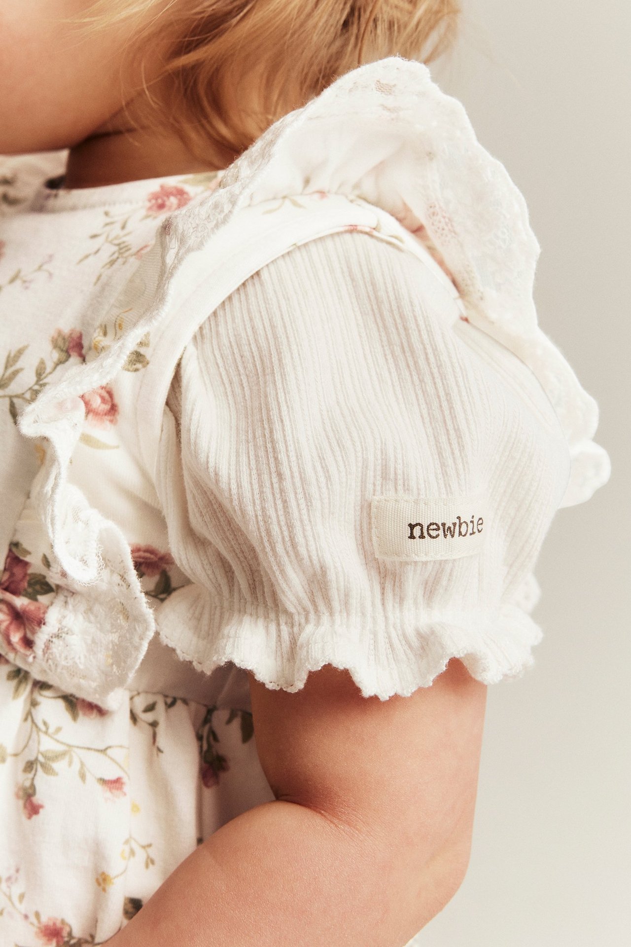 Close-up of a baby wearing an off-white floral sleeveless dress with lace over a ribbed off-white top.