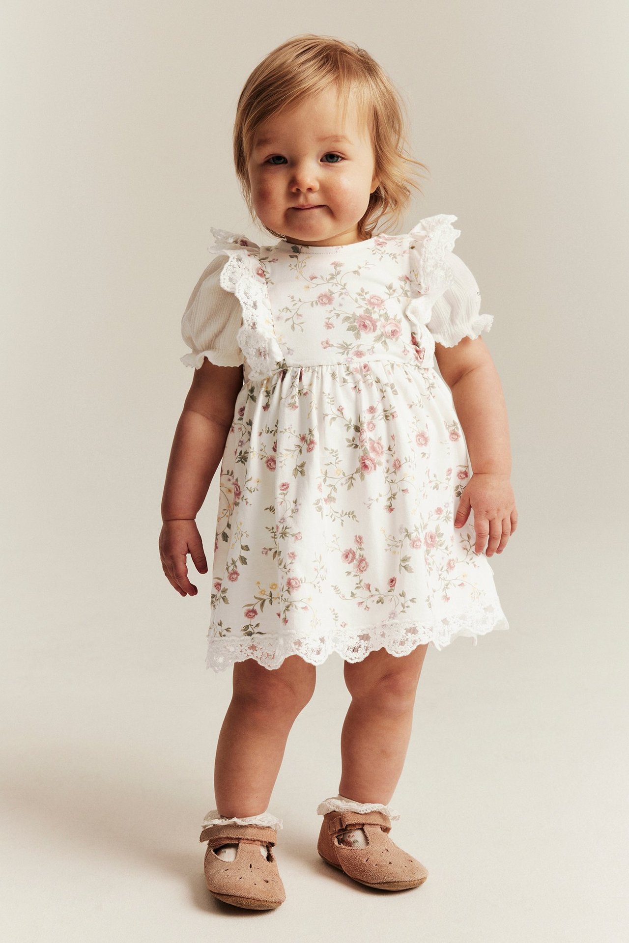 Front view: baby wears off-white floral jersey dress with lace, ribbed top, brown T-bar shoes.