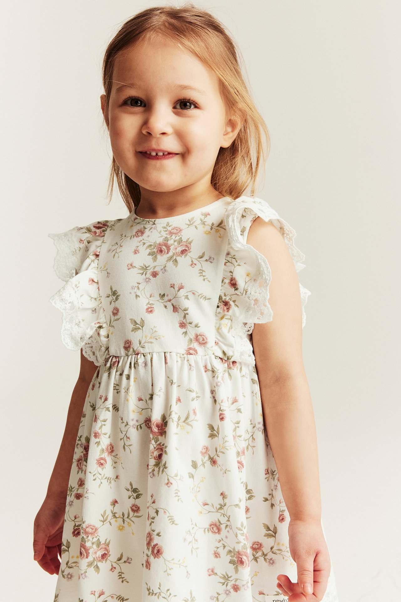 Front view: kid in an offwhite sleeveless floral jersey dress with lace ruffles on shoulders and hem.