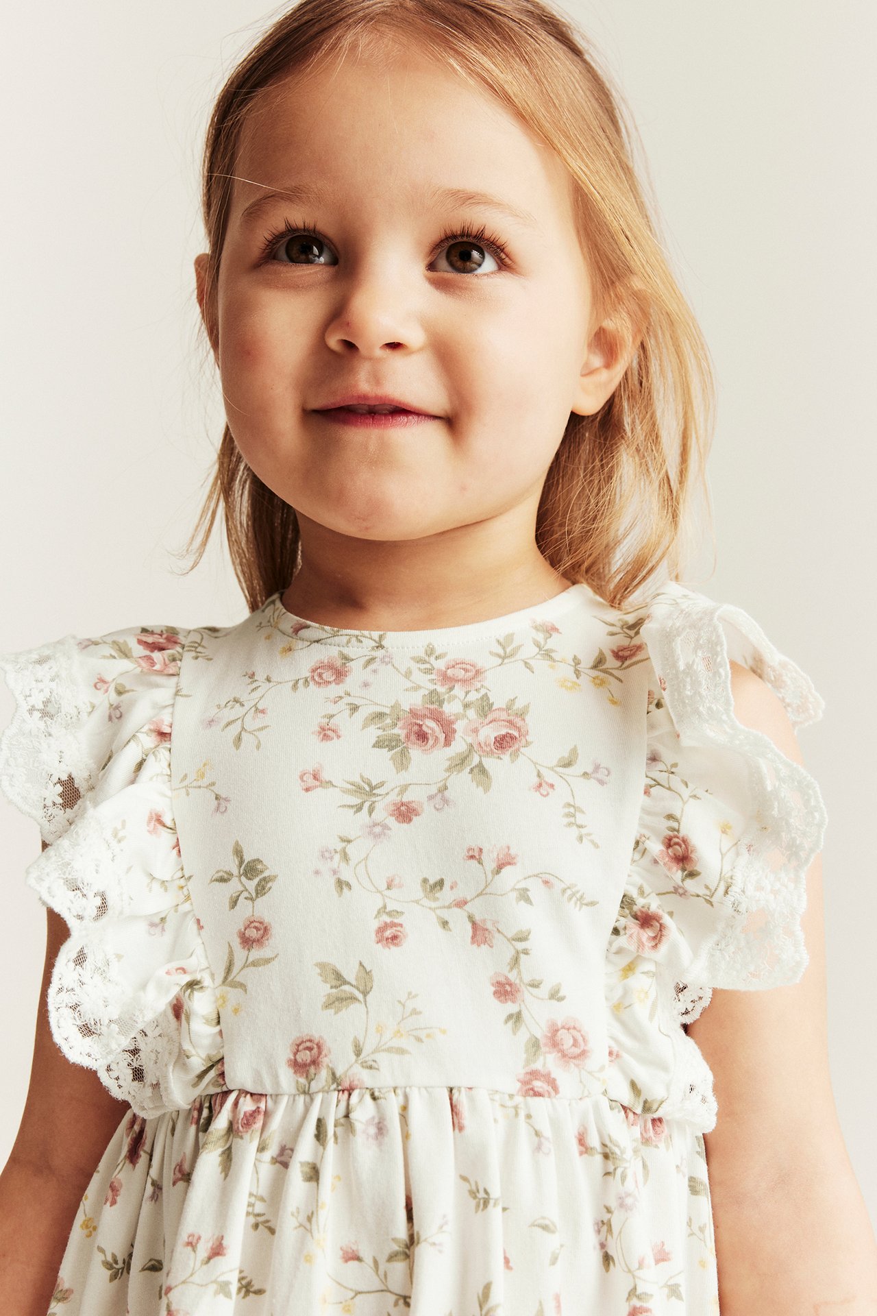 Close-up front view: kid wears offwhite floral jersey dress with pink roses and lace shoulder ruffles.