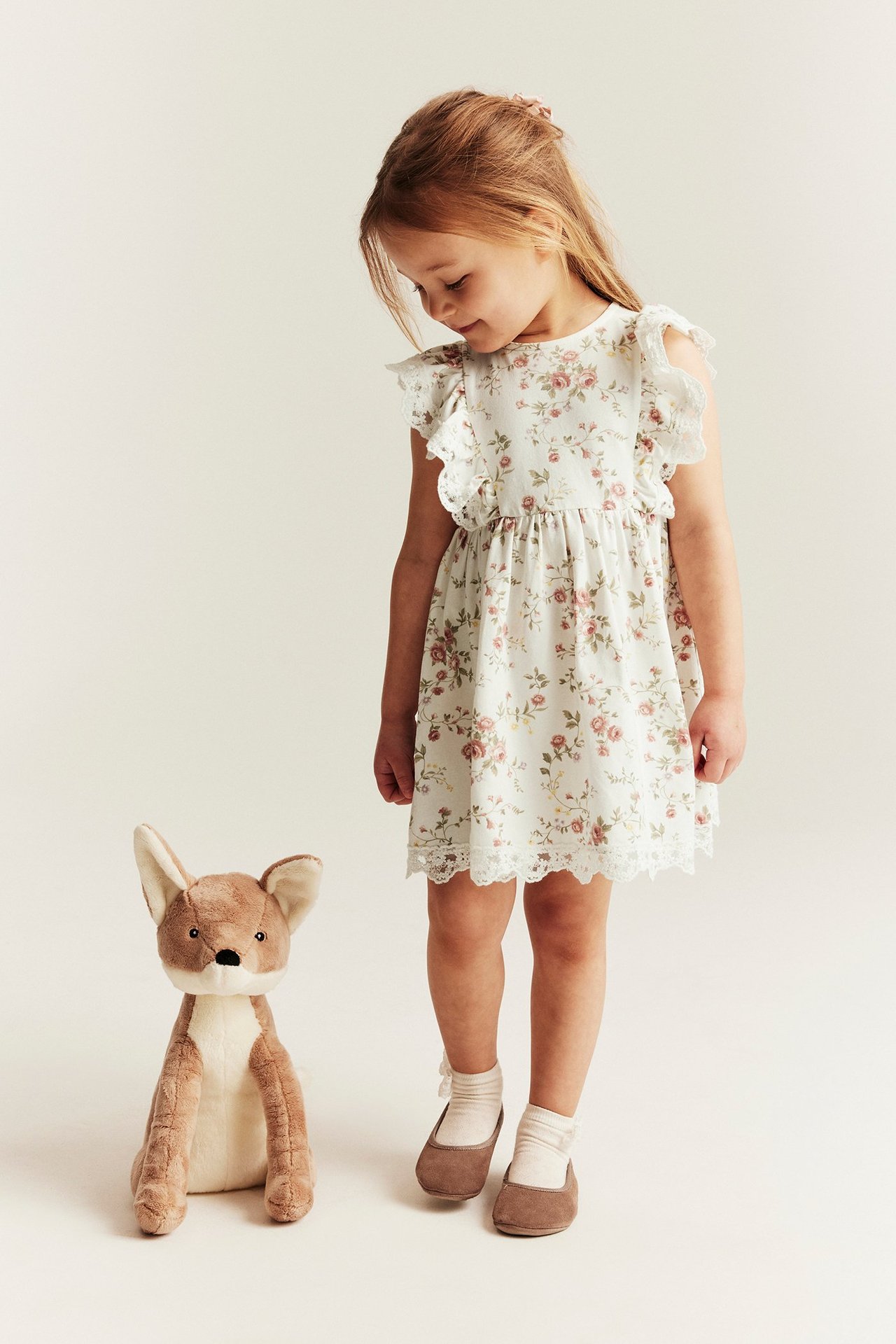 Front view of a kid in off-white floral sleeveless dress with ruffled shoulders and lace, next to a plush fox.
