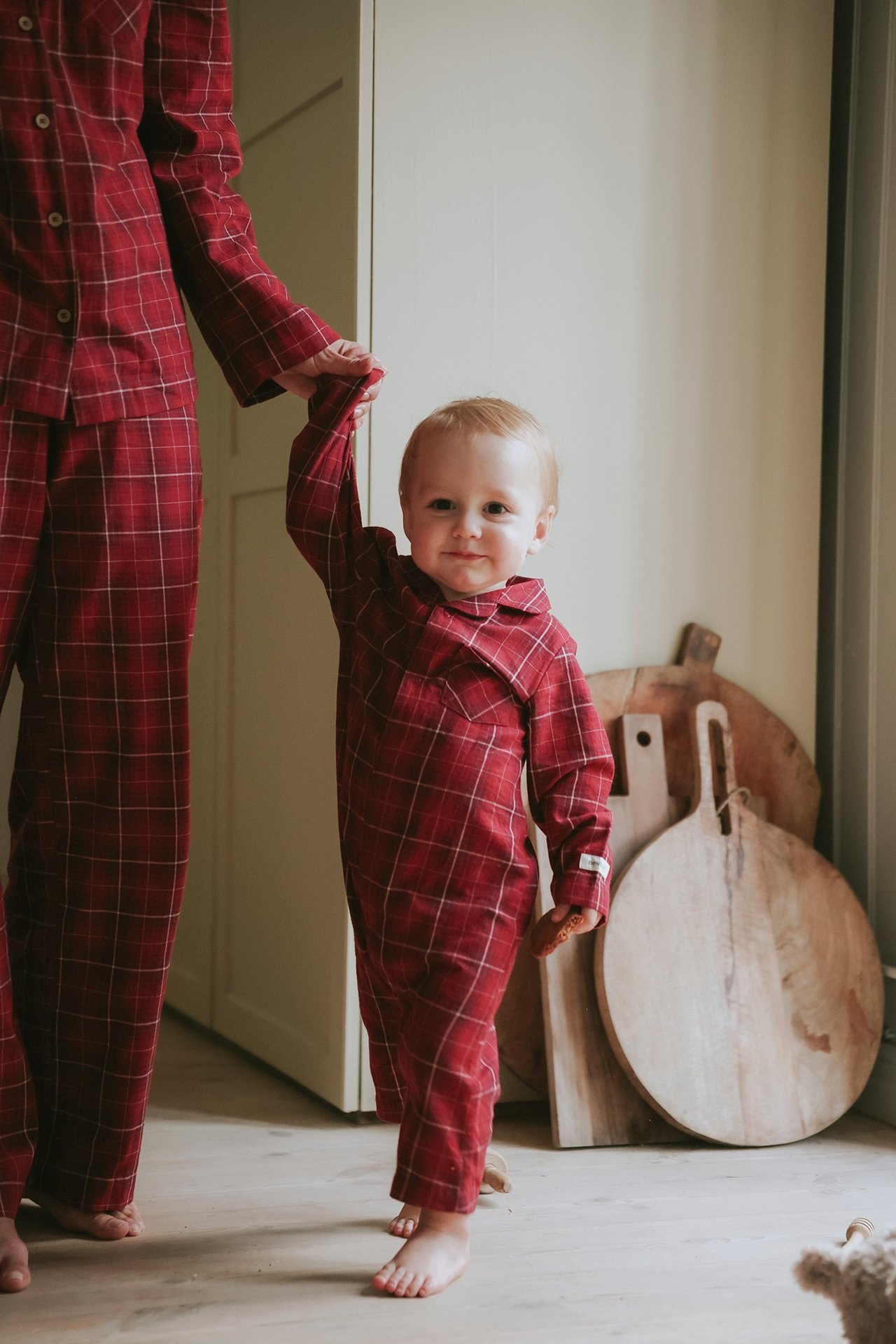 Front view: baby in red plaid flannel pyjama with snap buttons, holding an adult's hand.