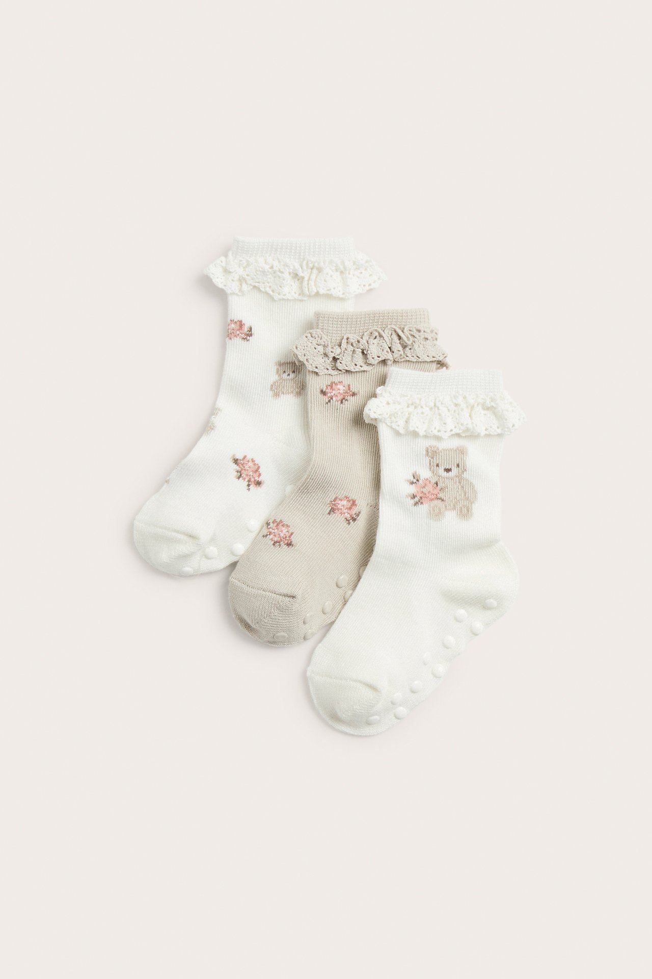 Three pairs of baby socks: off-white and beige with bear, rose motifs, lace cuffs, and anti-slip soles.