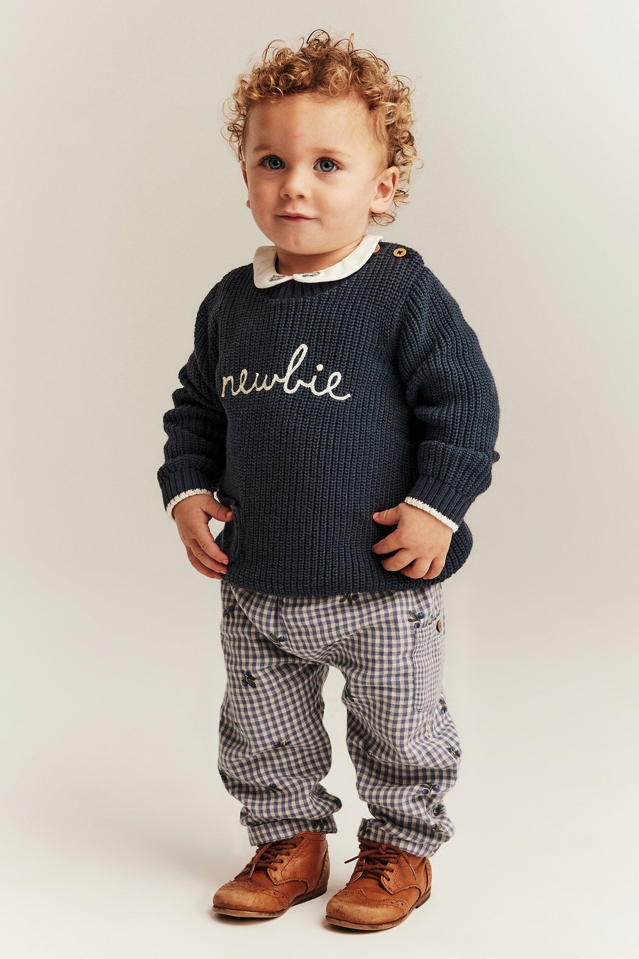 Front view: Baby wears a dark blue knitted 'newbie' sweater with white collar, checked trousers, and brown boots.