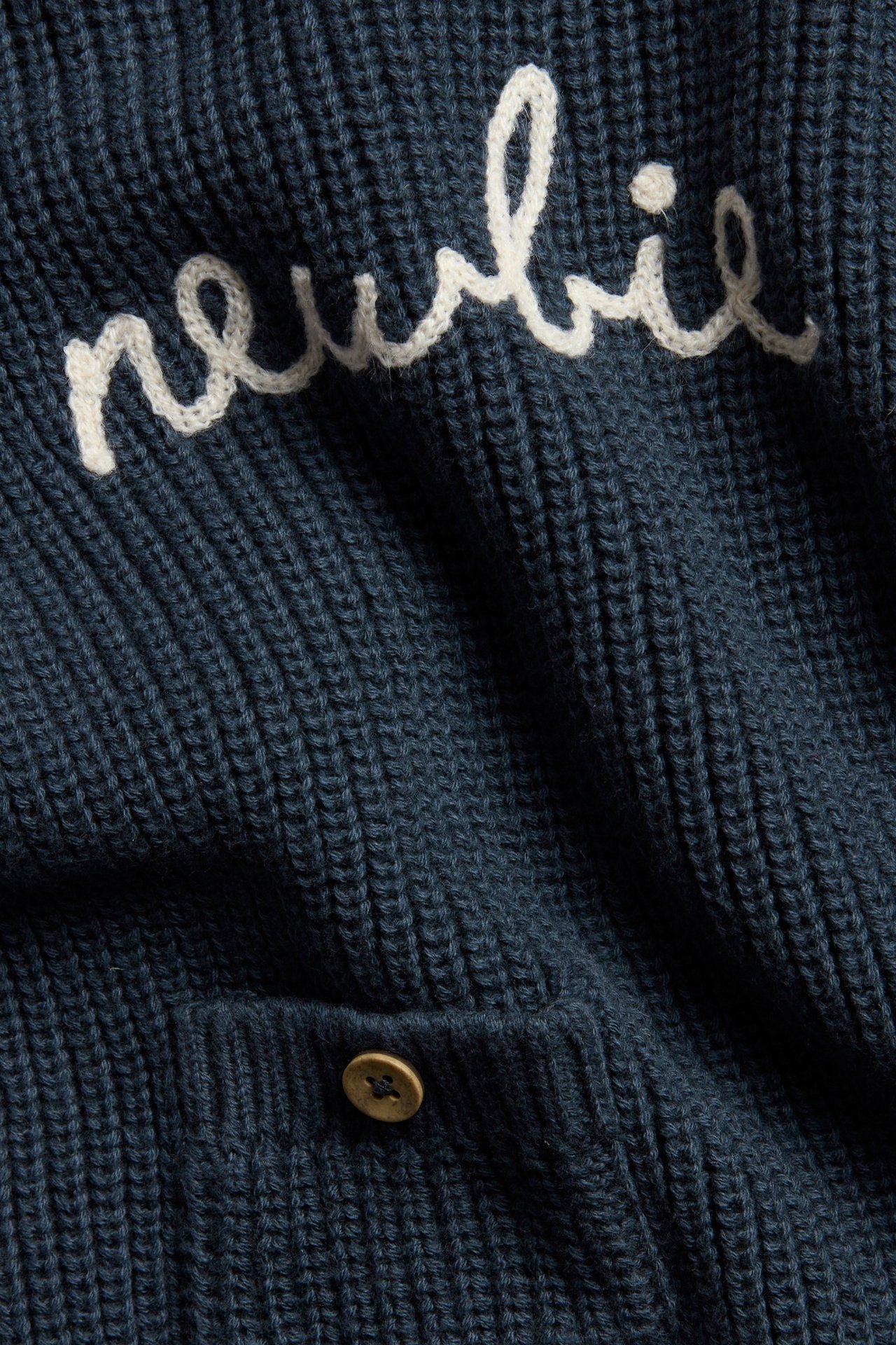 Close-up of a dark blue knitted cotton sweater with white 'Newbie' embroidery and a front pocket with a button.