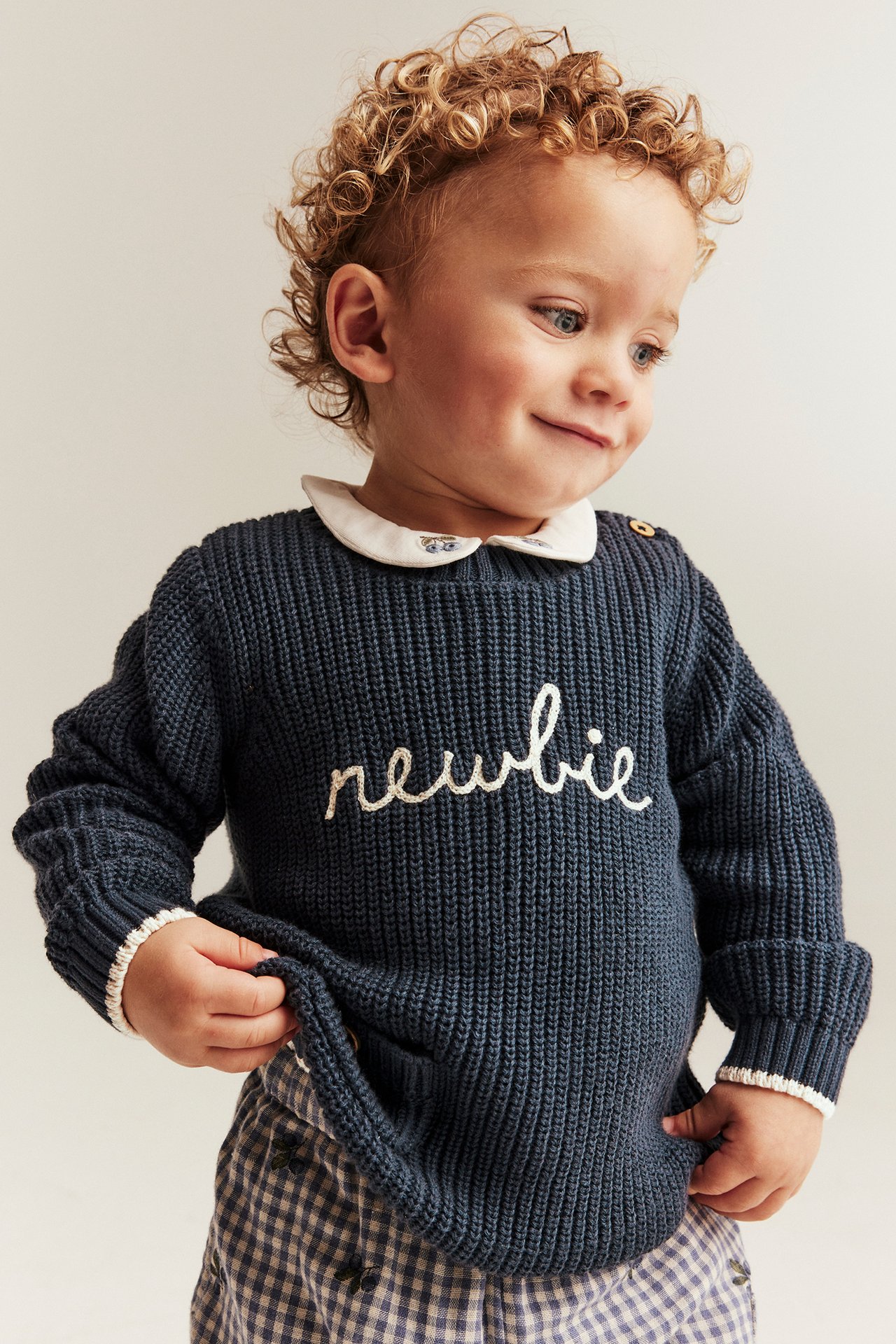 Front view of a baby boy wearing dark blue knitted sweater with "newbie" embroidery, white collar, and checkered pants.