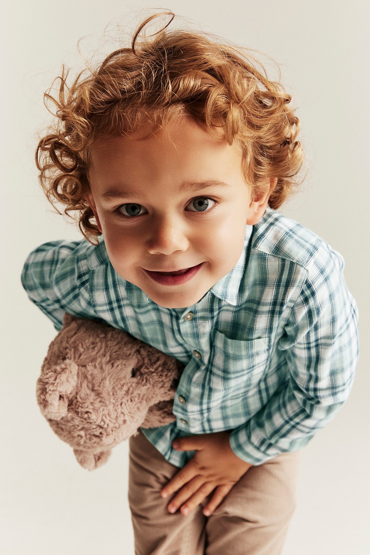 Kind in hellgrünem kariertem Baumwollhemd, beiger Hose, hält einen braunen Teddybär.