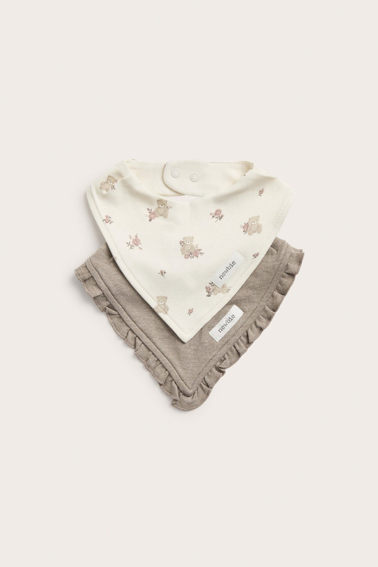 Zwei Baby-Bandana-Lätzchen: eines cremefarben mit Teddybär- und Blumenmuster, eines einfarbig braun mit Rüschen.