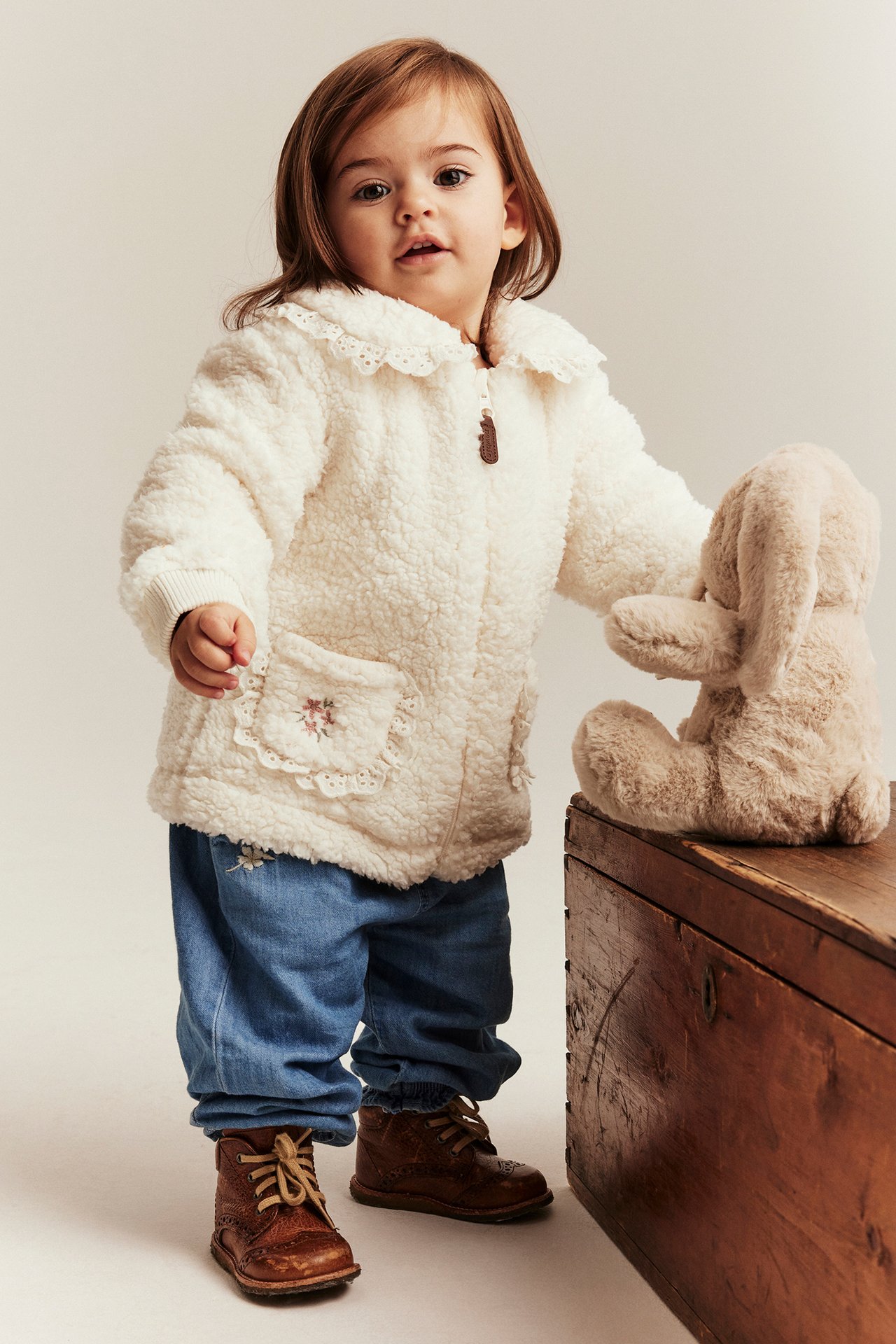 Front view: Baby girl in an offwhite pile jacket with lace details, blue denim trousers, and brown boots.