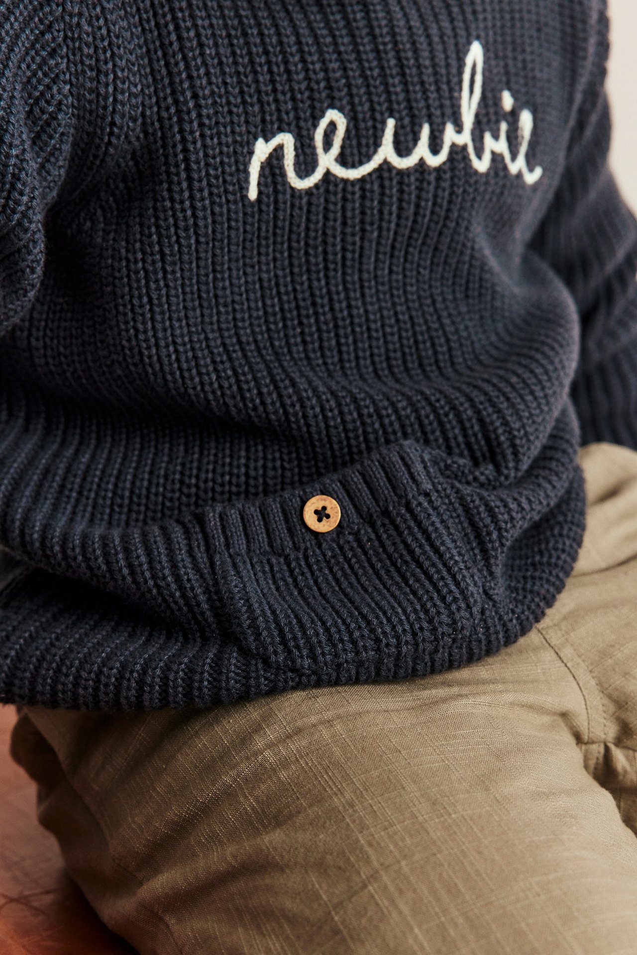 Dunkelblauer Newbie-Strickpullover für Kinder mit weißer Stickerei, Holzknopf und beiger Hose.