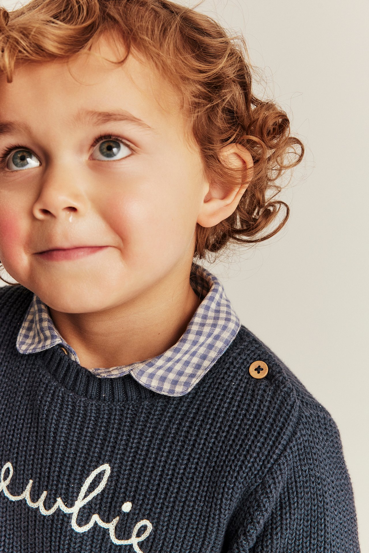 Close-up: Kid wears a dark blue knitted Newbie sweater with shoulder button over a gingham shirt.