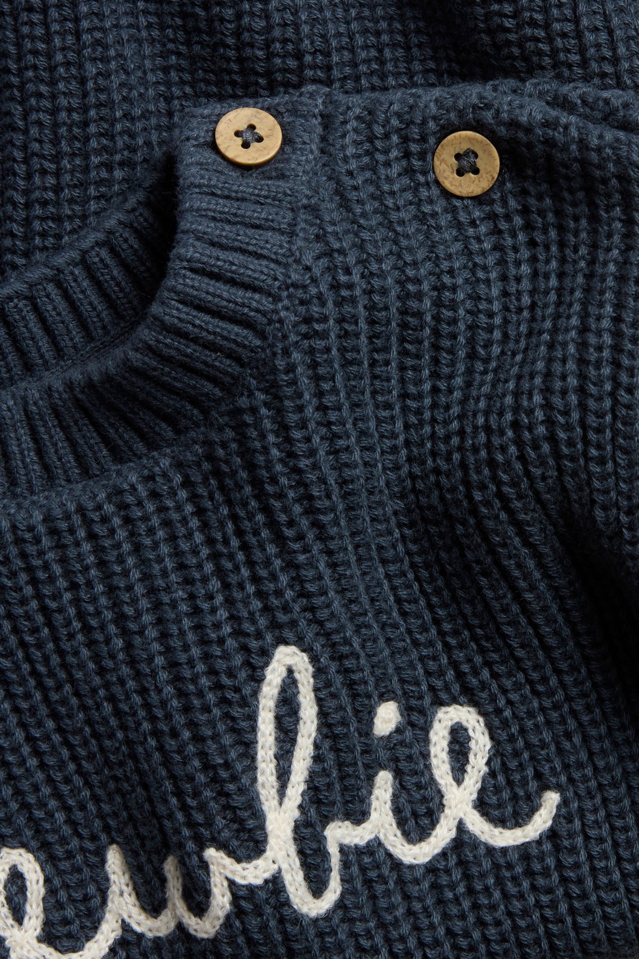 Close-up of a dark blue knitted cotton sweater with white 'Newbie' embroidery and two wooden buttons on the shoulder.