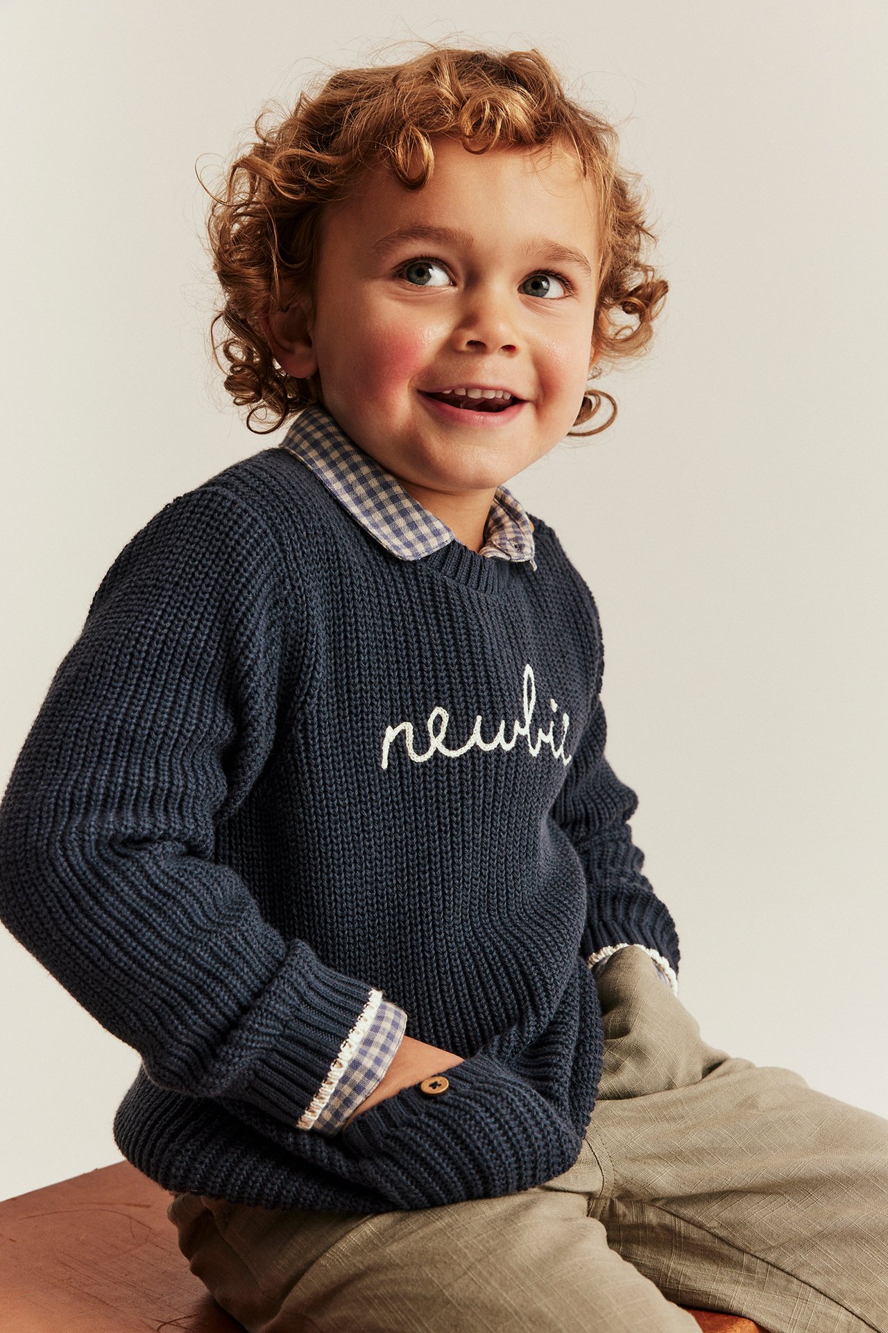 Front view of a kid wearing a dark blue knitted Newbie sweater over a checkered shirt and khaki trousers.