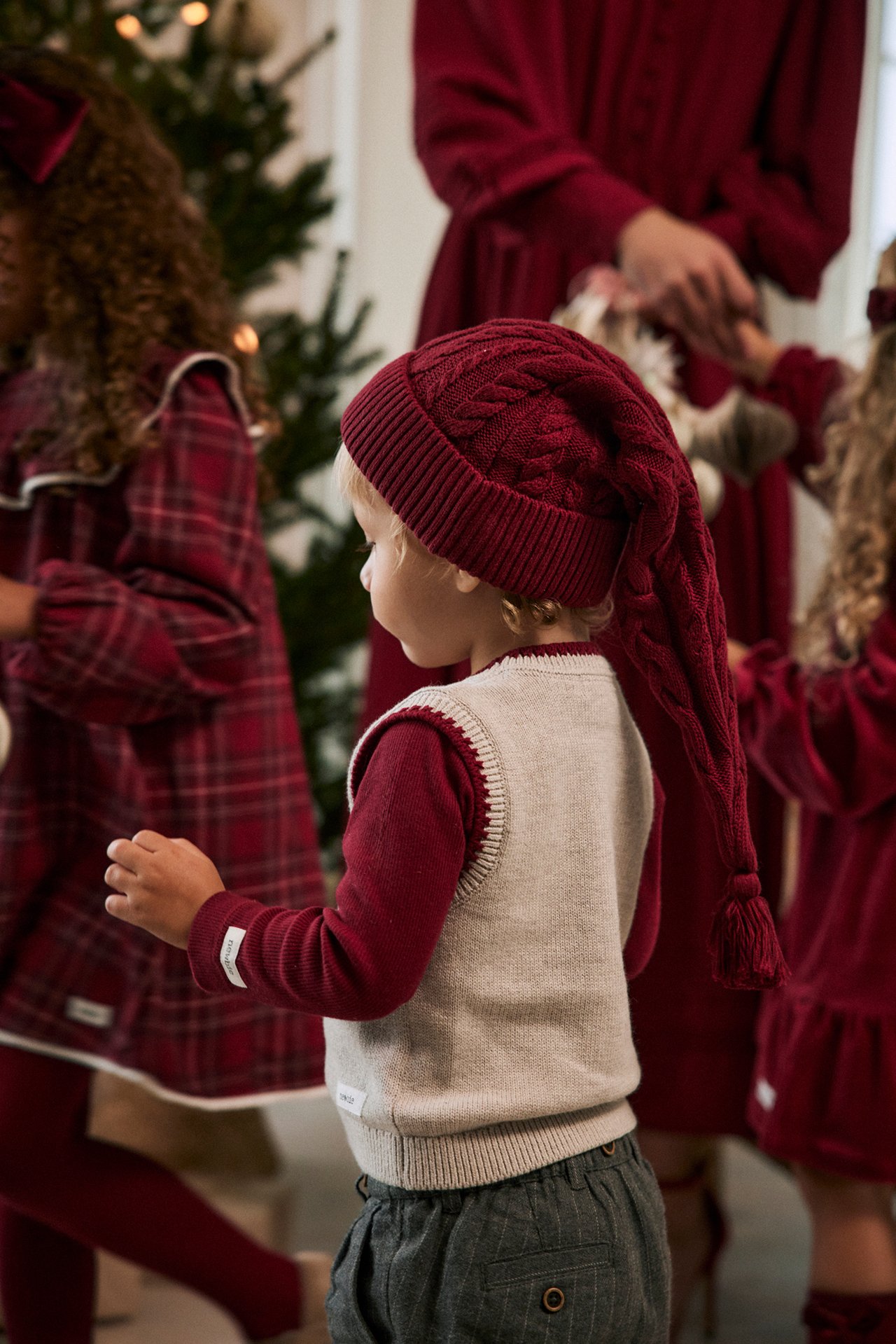 Back view of a baby boy wearing a red cable-knitted long Santa hat, red top, light beige vest, and grey trousers.