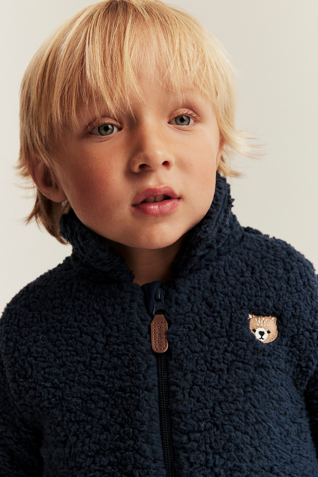 Front view of a kid wearing a dark blue pile jacket with a brown bear embroidery and a zipper.