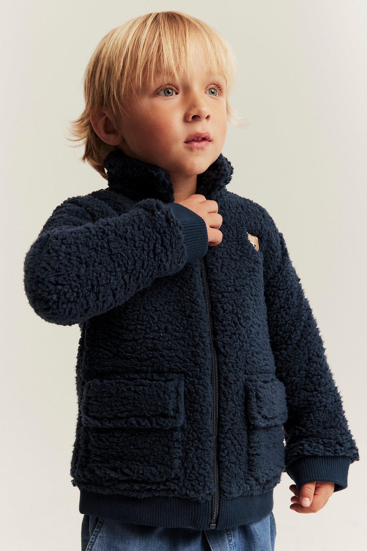 Kid wears a dark blue fleece jacket with bear embroidery, a full zipper, and front patch pockets.