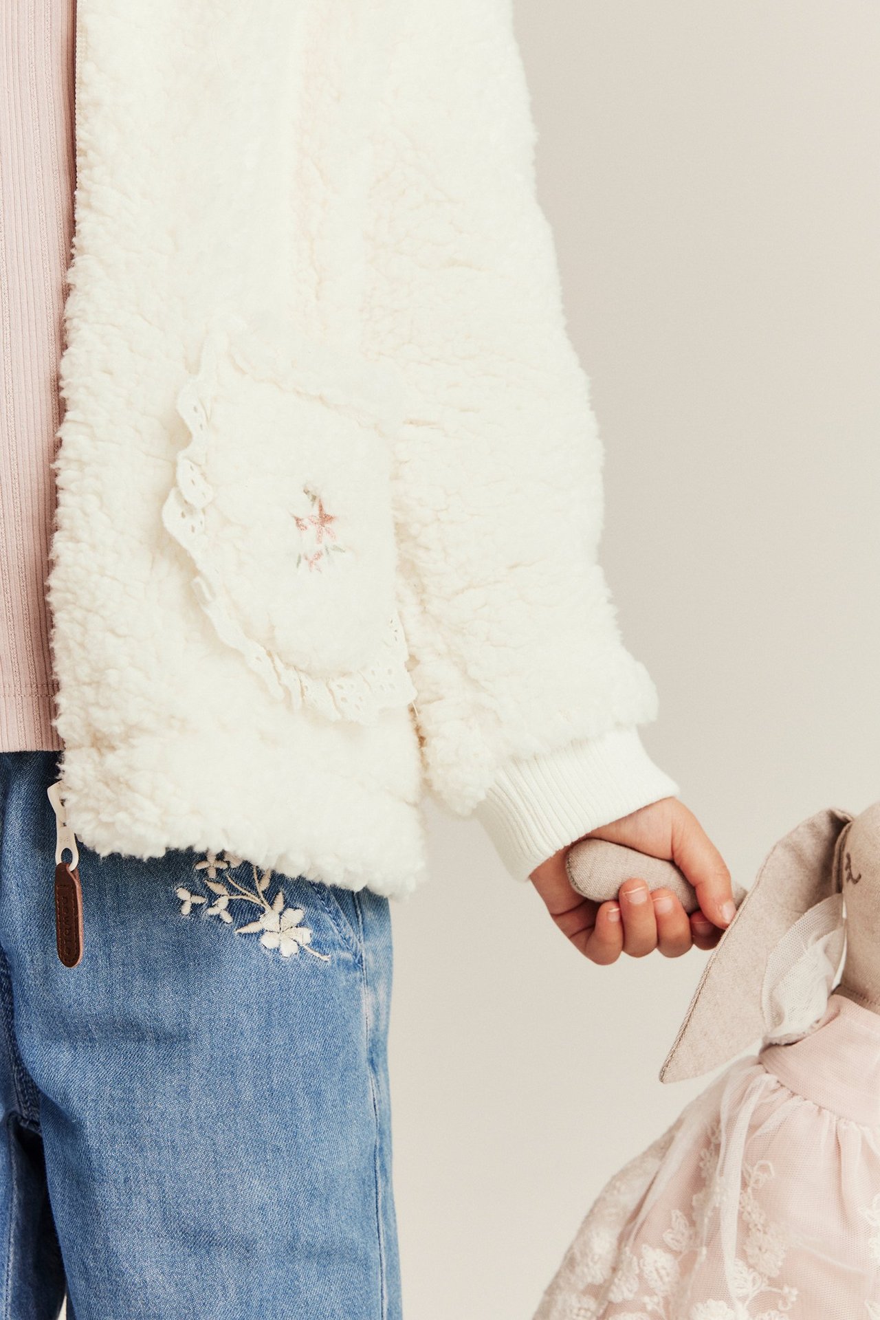 Kind trägt cremeweiße Plüschjacke mit Spitze und Stickerei, pinkes Oberteil, blaue bestickte Jeans, hält Hasenpuppe.