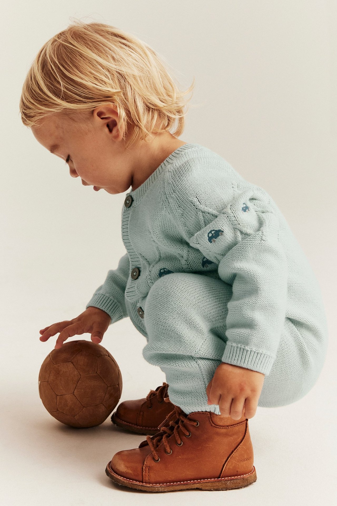 Seitenansicht: Baby in helltürkisem, strukturgestricktem Baumwoll-Jumpsuit mit Auto-Stickerei und braunen Stiefeln.