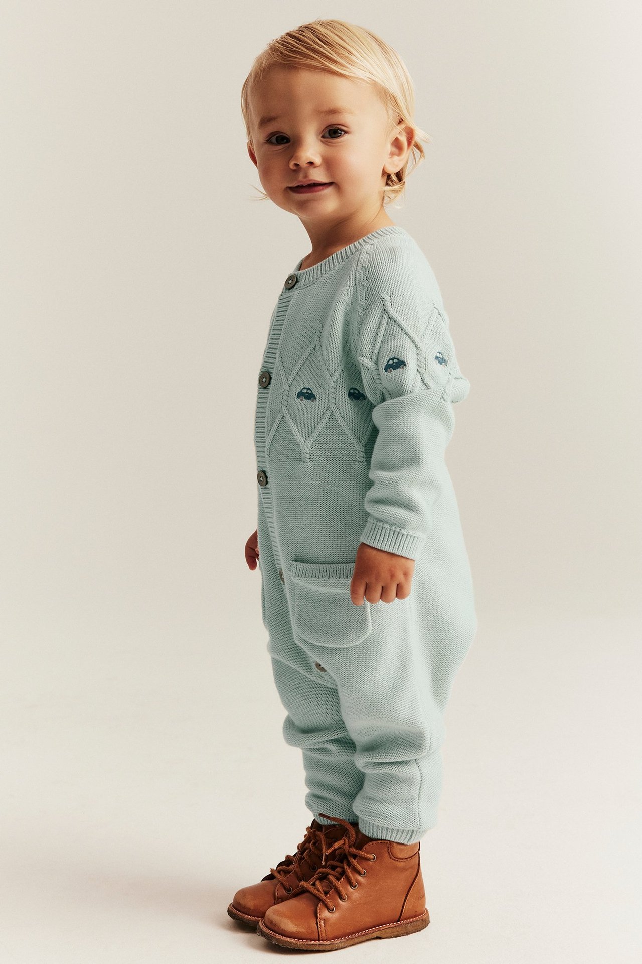Seitenansicht eines Kindes in helltürkisem gestricktem Baumwoll-Jumpsuit mit Auto-Stickerei und braunen Stiefeln.
