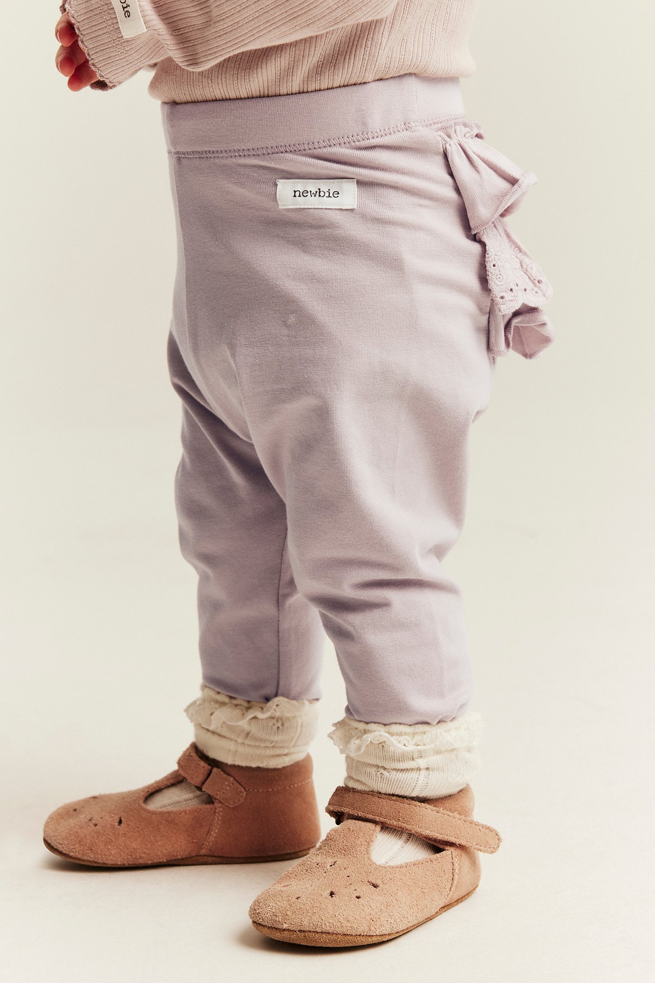 Side view of a baby wearing lavender pants with ruffles and a white 'newbie' label, white ruffled socks, and brown suede shoes.