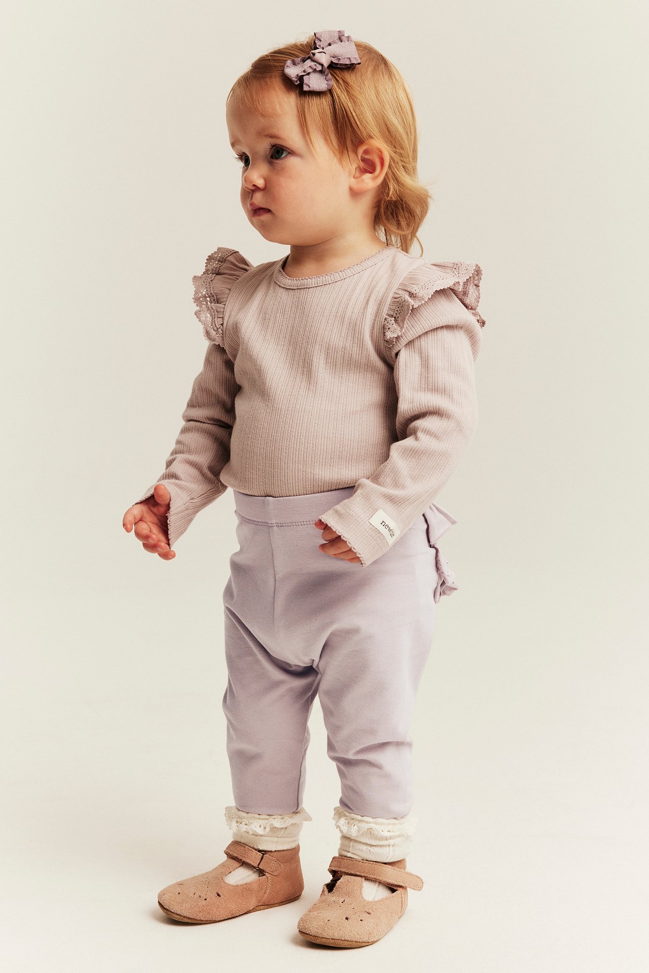 Side view: baby wears a mauve ribbed top, lavender leggings, brown shoes, and purple bow.