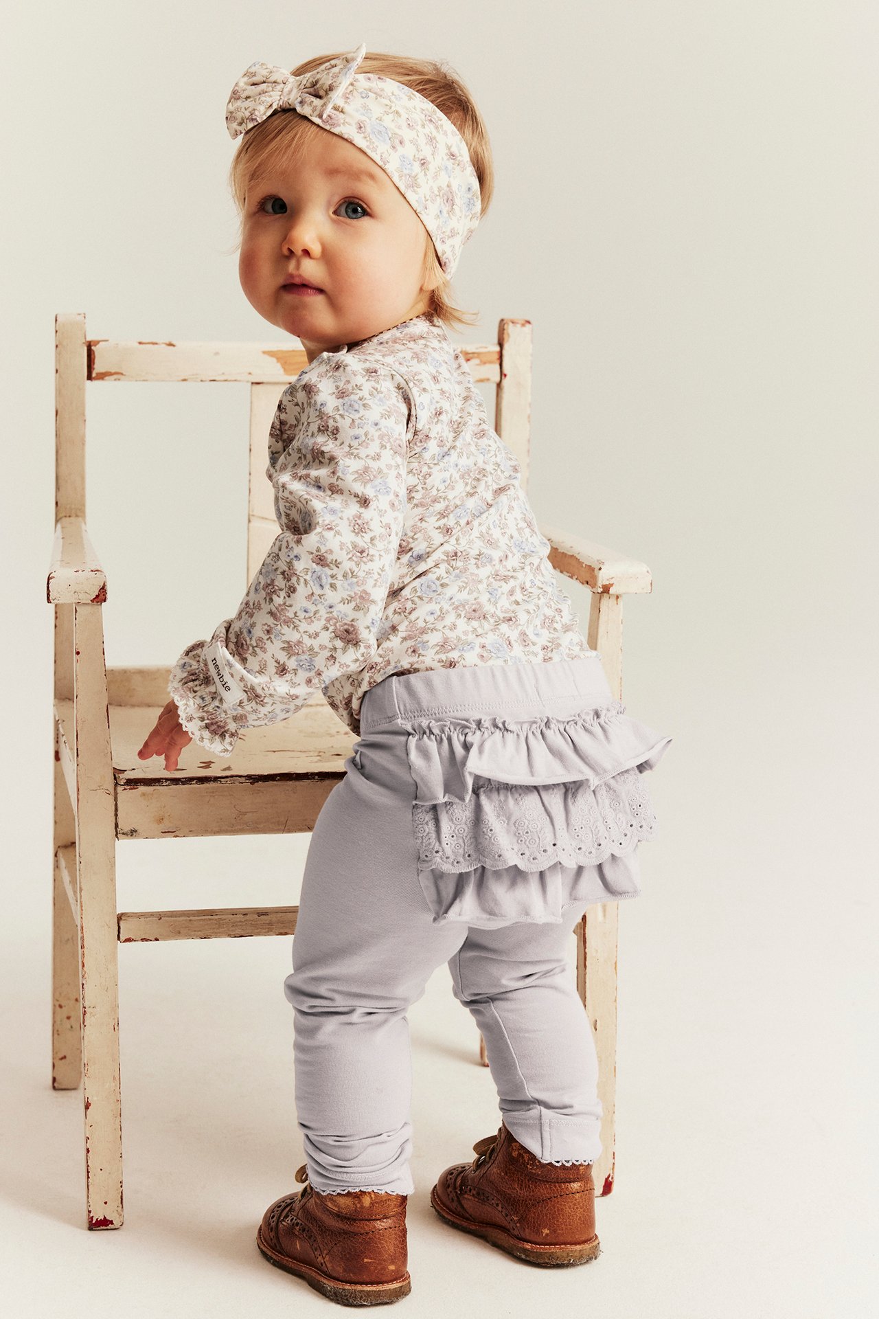 Back-side view of a baby in light lilac ruffled lace leggings, floral top, matching headband, and brown shoes.