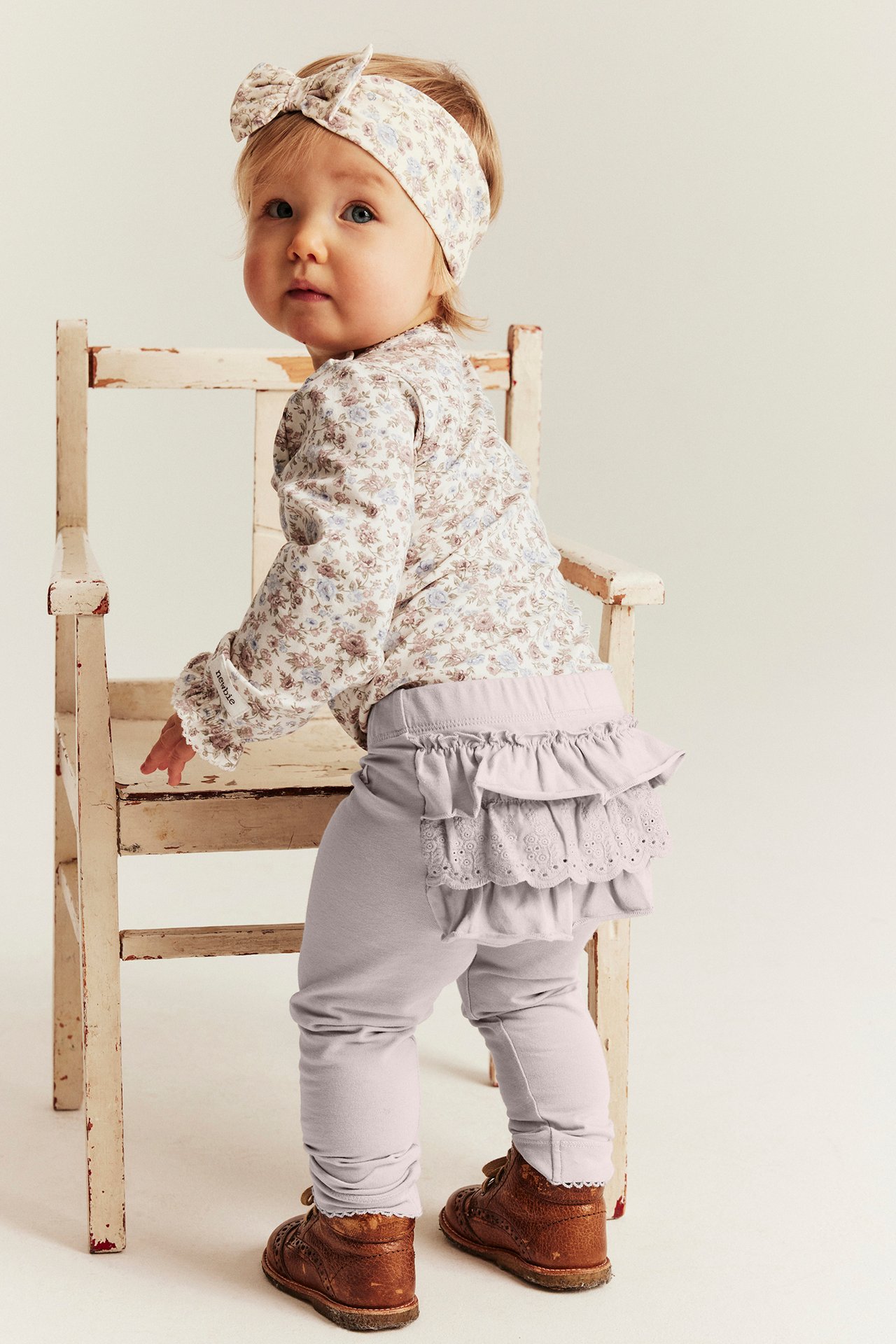 Rückansicht: Baby in helllila Rüschen-Leggings, Blümchen-Top, passendem Stirnband und braunen Schuhen.