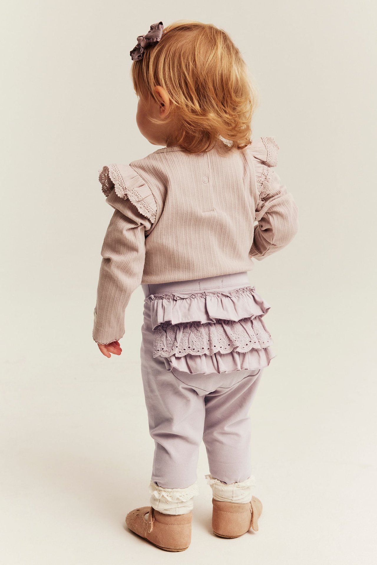Rückenansicht eines Babys in lila Rüschenleggings, einem rosa Rippentop und beigen Schuhen.