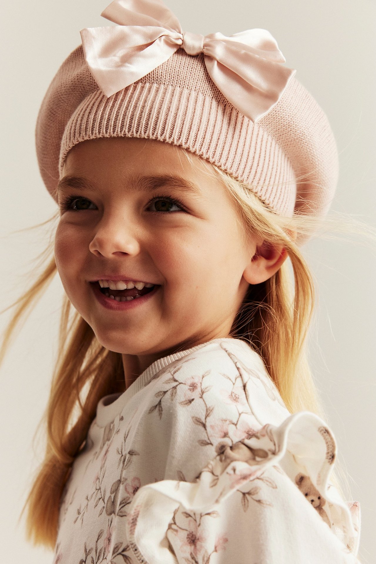 Nahaufnahme eines Mädchens in cremeweißem Sweatshirt mit Blumen- und Bärendruck und Rüschenschultern, plus rosa Strickbarett.