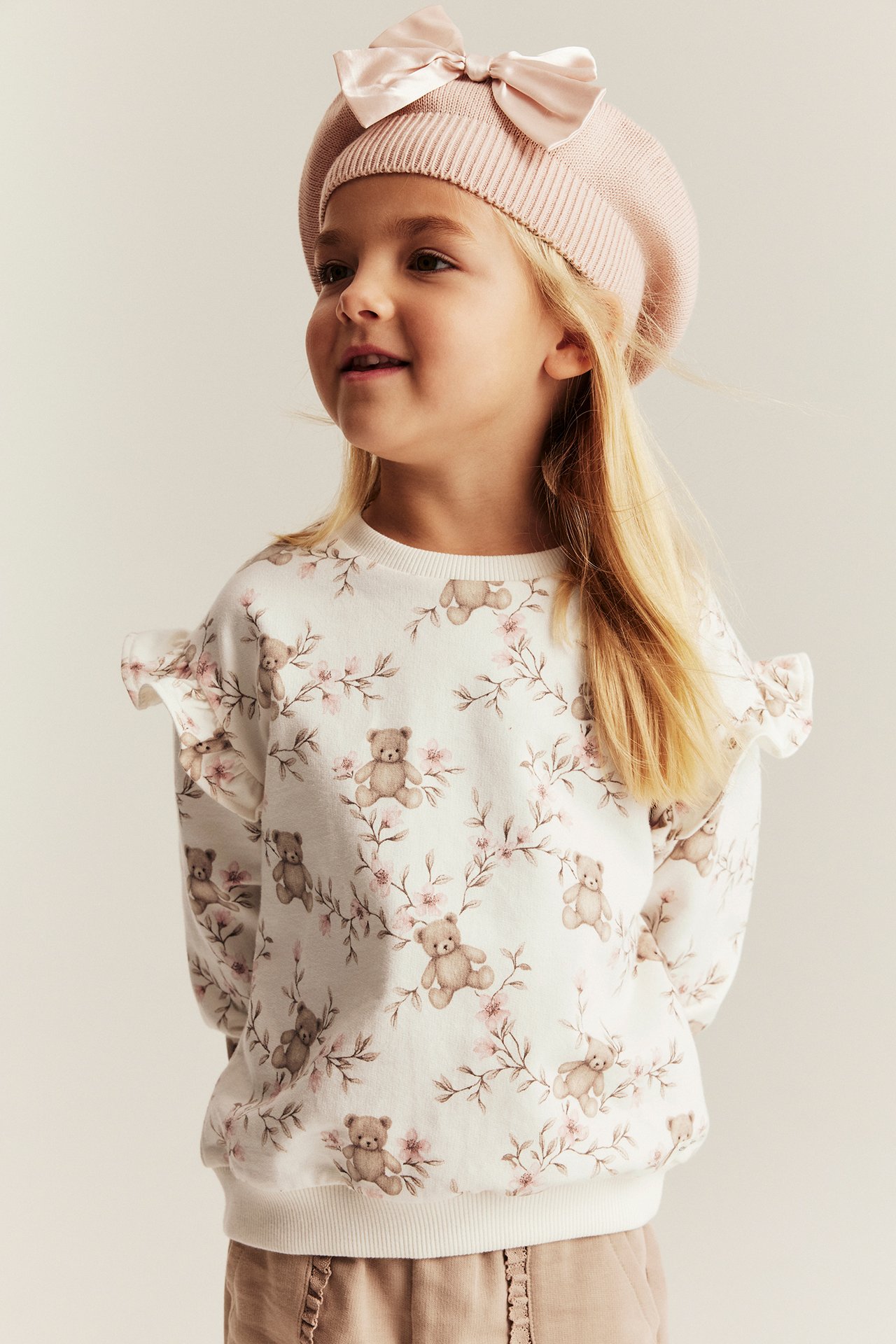 Vorderansicht: Kind trägt cremefarbenes Baumwoll-Sweatshirt mit Bärendruck, hellbraune Hose und rosa Baskenmütze.