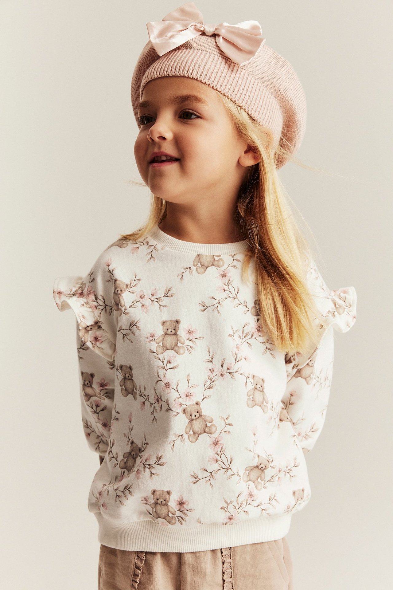 Vorderansicht: Kind trägt cremefarbenes Baumwoll-Sweatshirt mit Bärendruck, hellbraune Hose und rosa Baskenmütze.