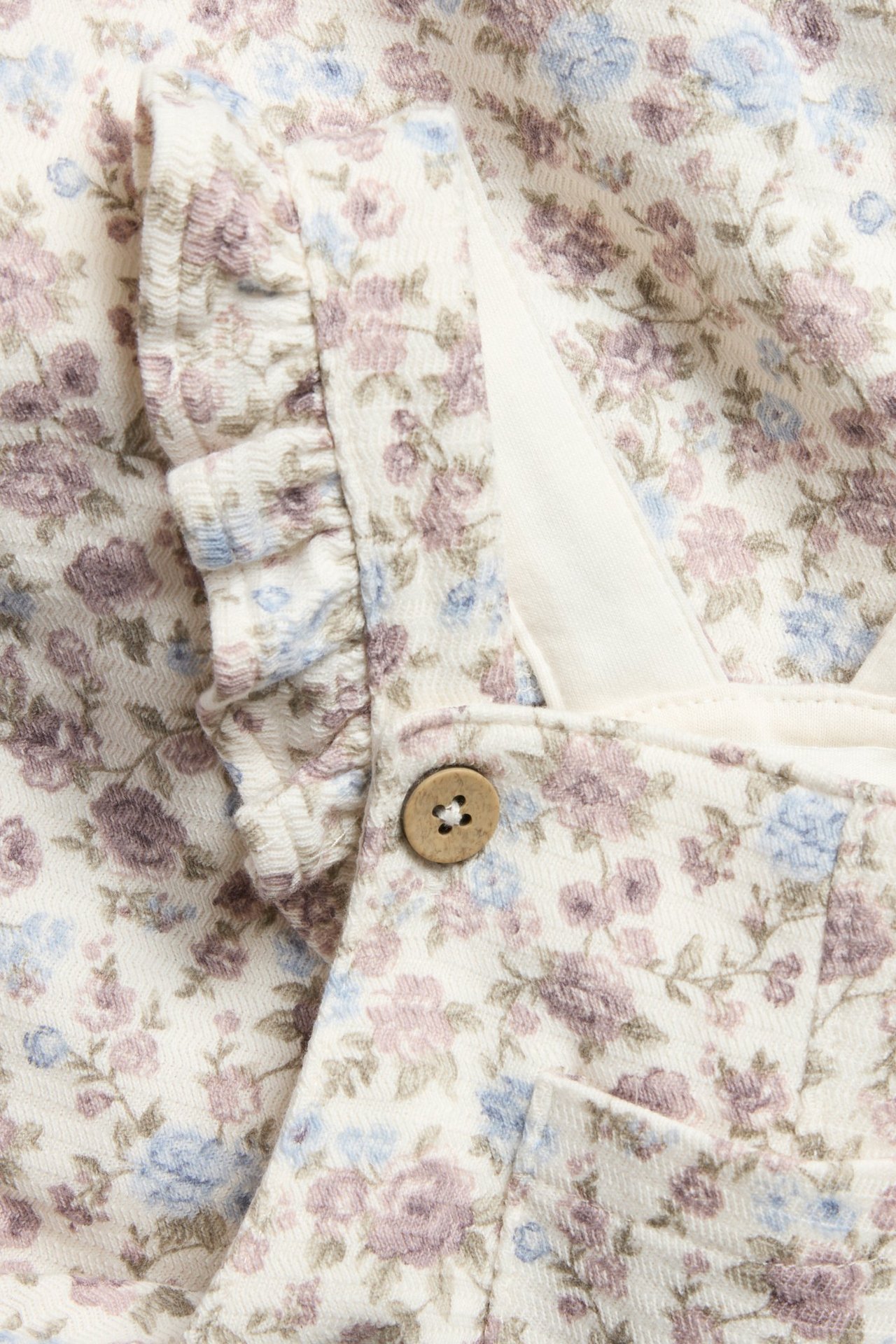 Nahaufnahme von off-white geblümten Jersey-Baby-Latzhosen mit Rüschenträgern und einem Holzknopf.