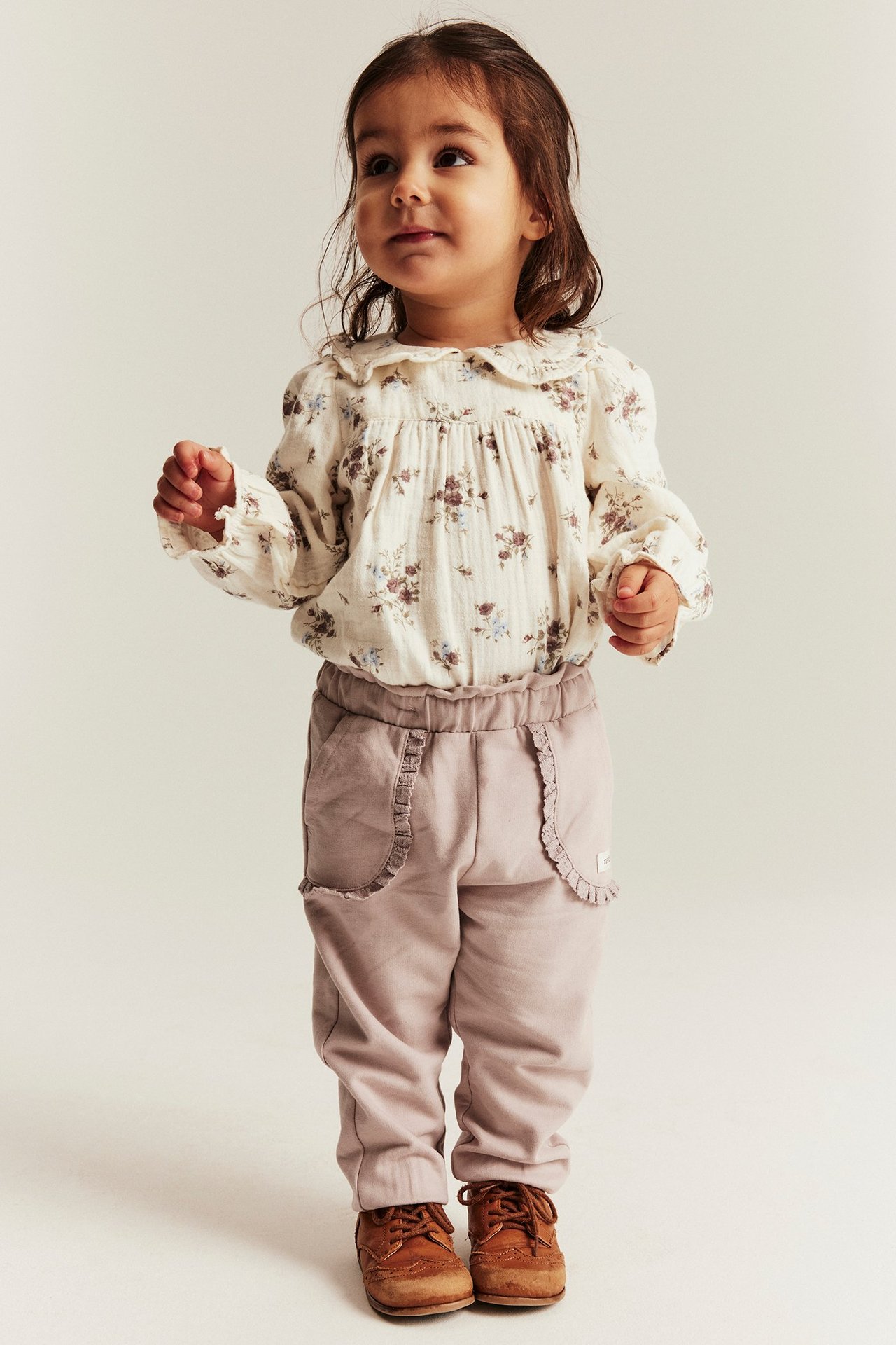 Front view: baby girl in off-white floral blouse body, light purple ruffled pants, and brown shoes.