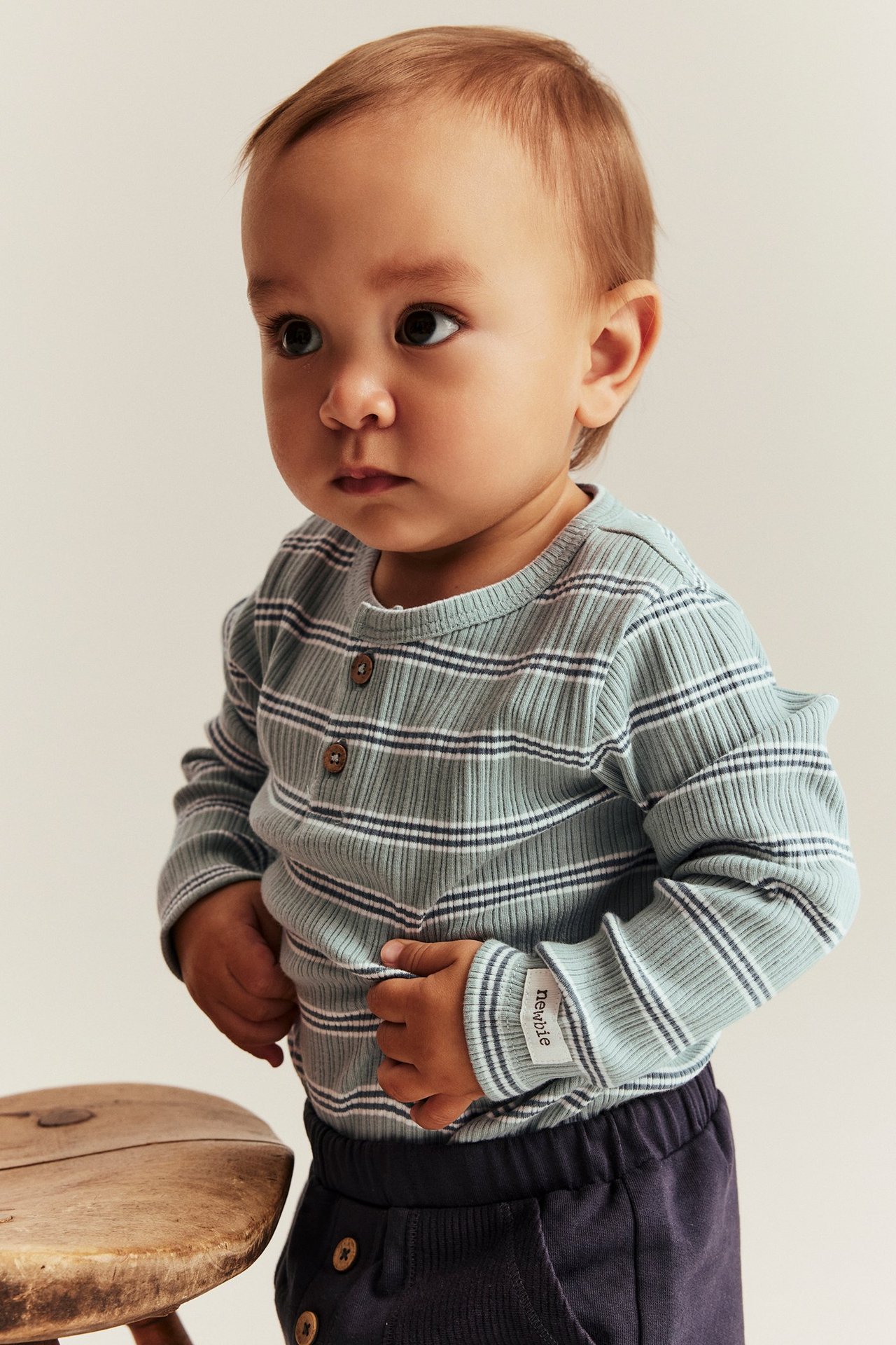 Vorderansicht, Baby trägt hellgrünen, gestreiften Ripp-Langarmbody mit zwei Knöpfen und dunkler blauer Hose.