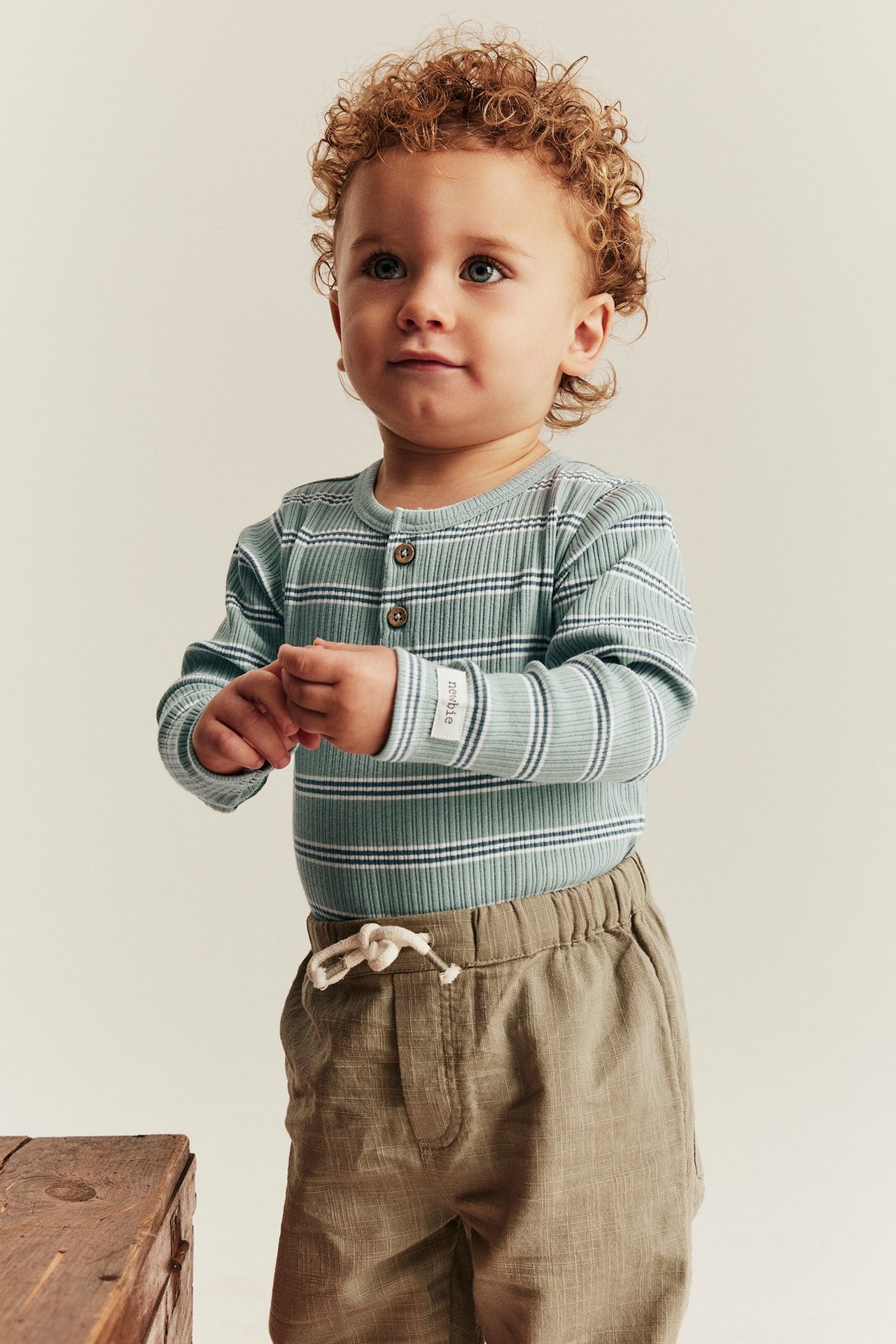 Vorderansicht: Baby trägt hellgrünen gestreiften gerippten Body mit Knöpfen und hellbraune Hose.