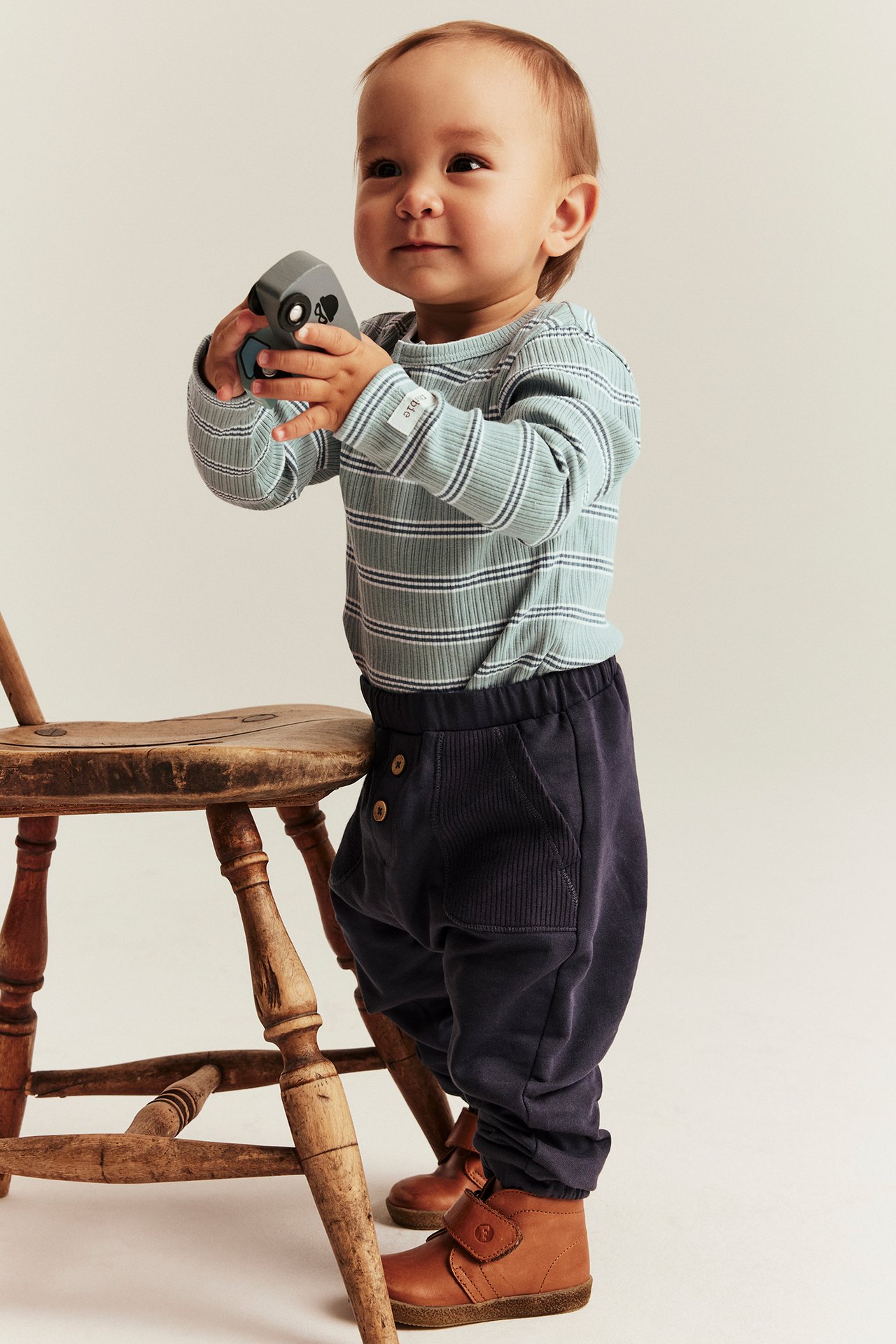 Front view: baby in a light green striped ribbed bodysuit, dark blue trousers, brown boots, holding a grey toy.