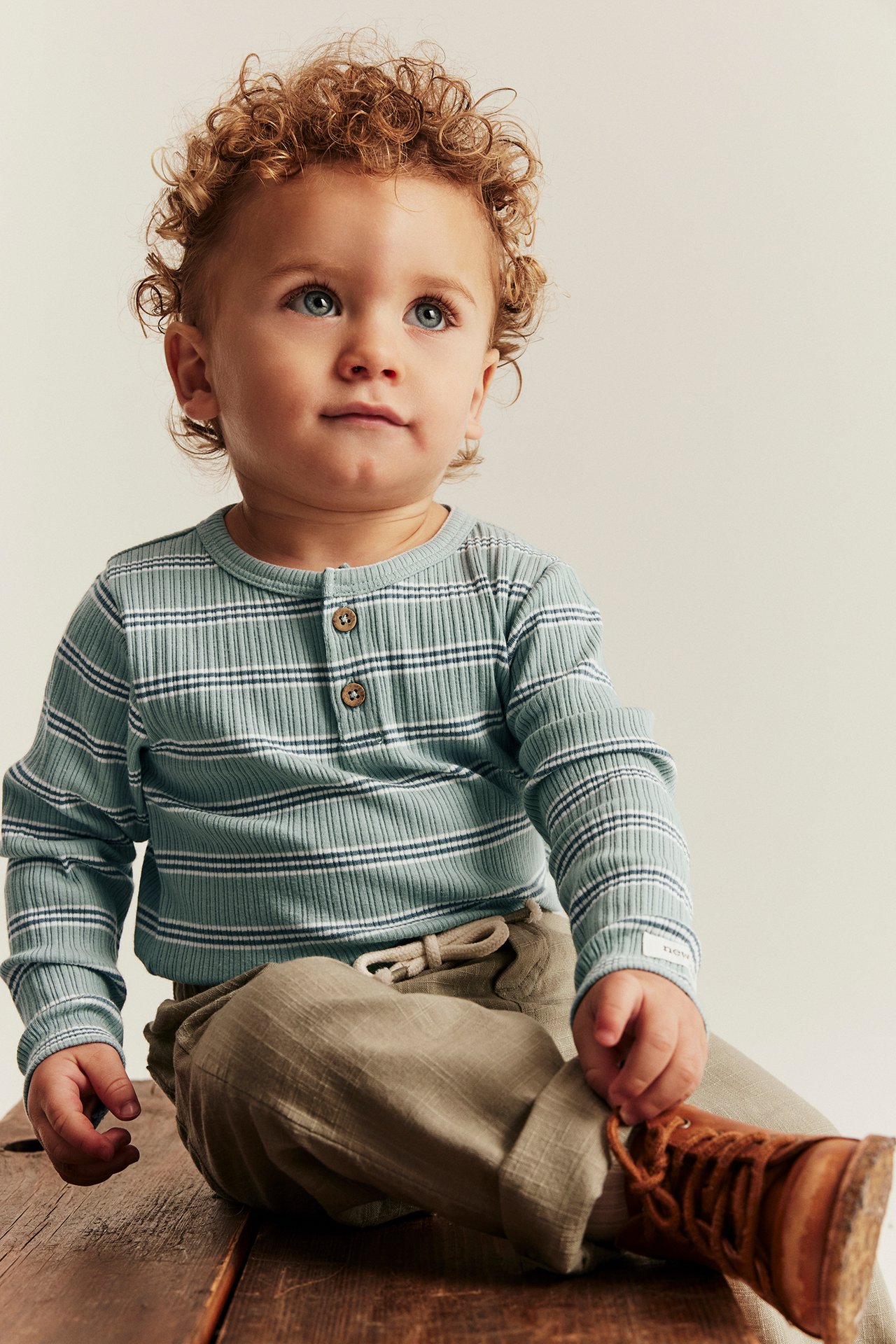 Front view of a baby in a light green striped ribbed long-sleeved body, light brown trousers, and brown laced boots.