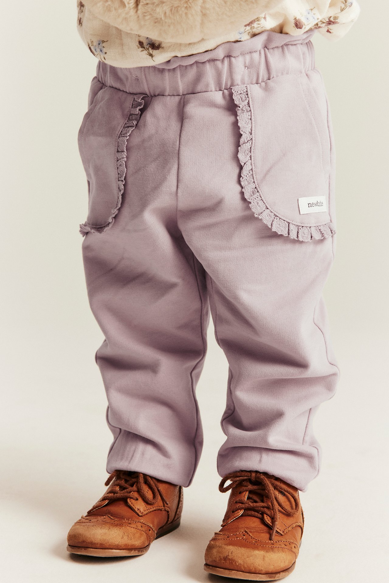 Front view of a baby in light lilac jogging pants with lace-trimmed pockets and brown shoes.