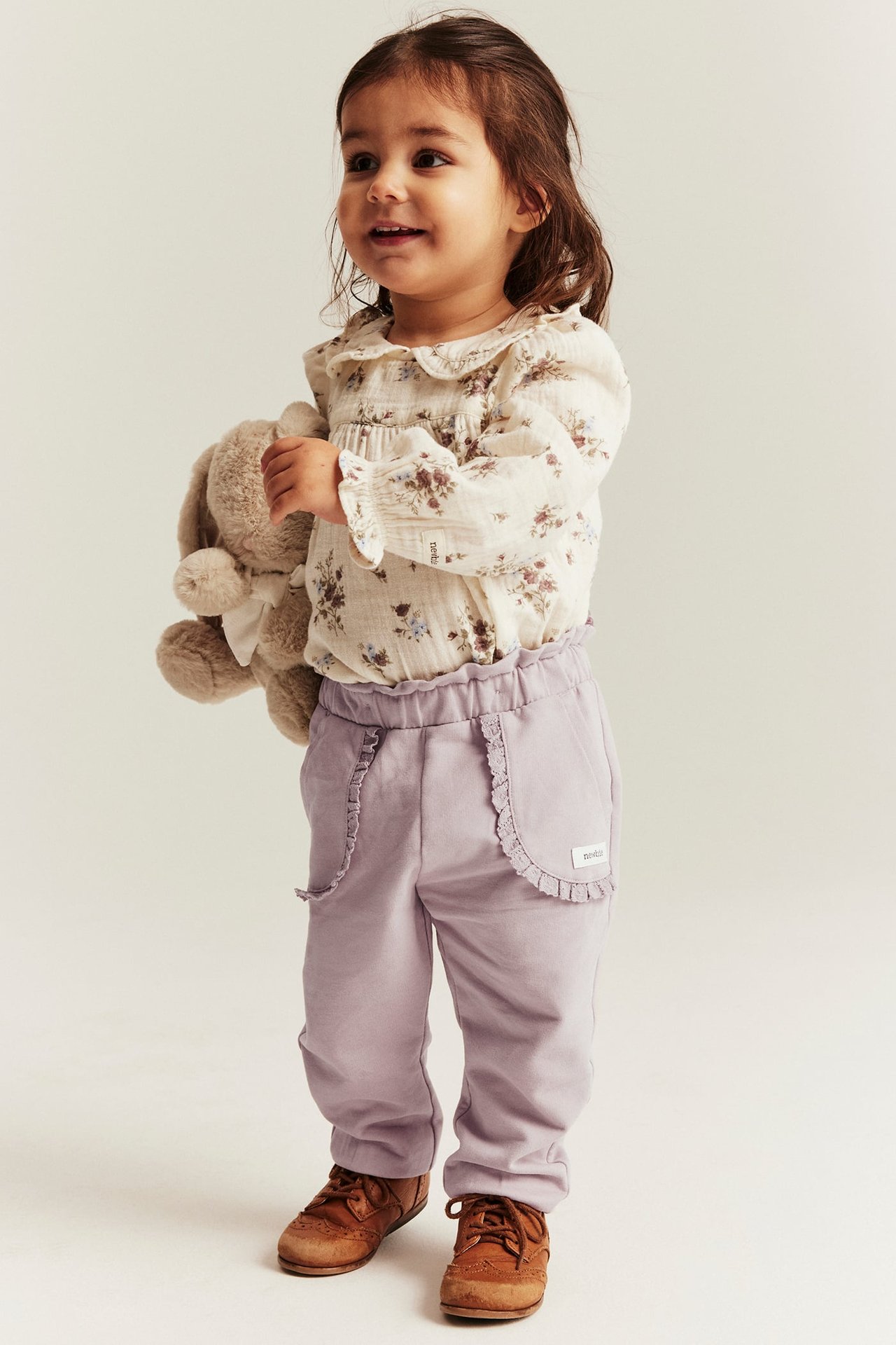 Baby girl in floral blouse, light purple pants with ruffled pockets, brown shoes, holding a beige bunny.