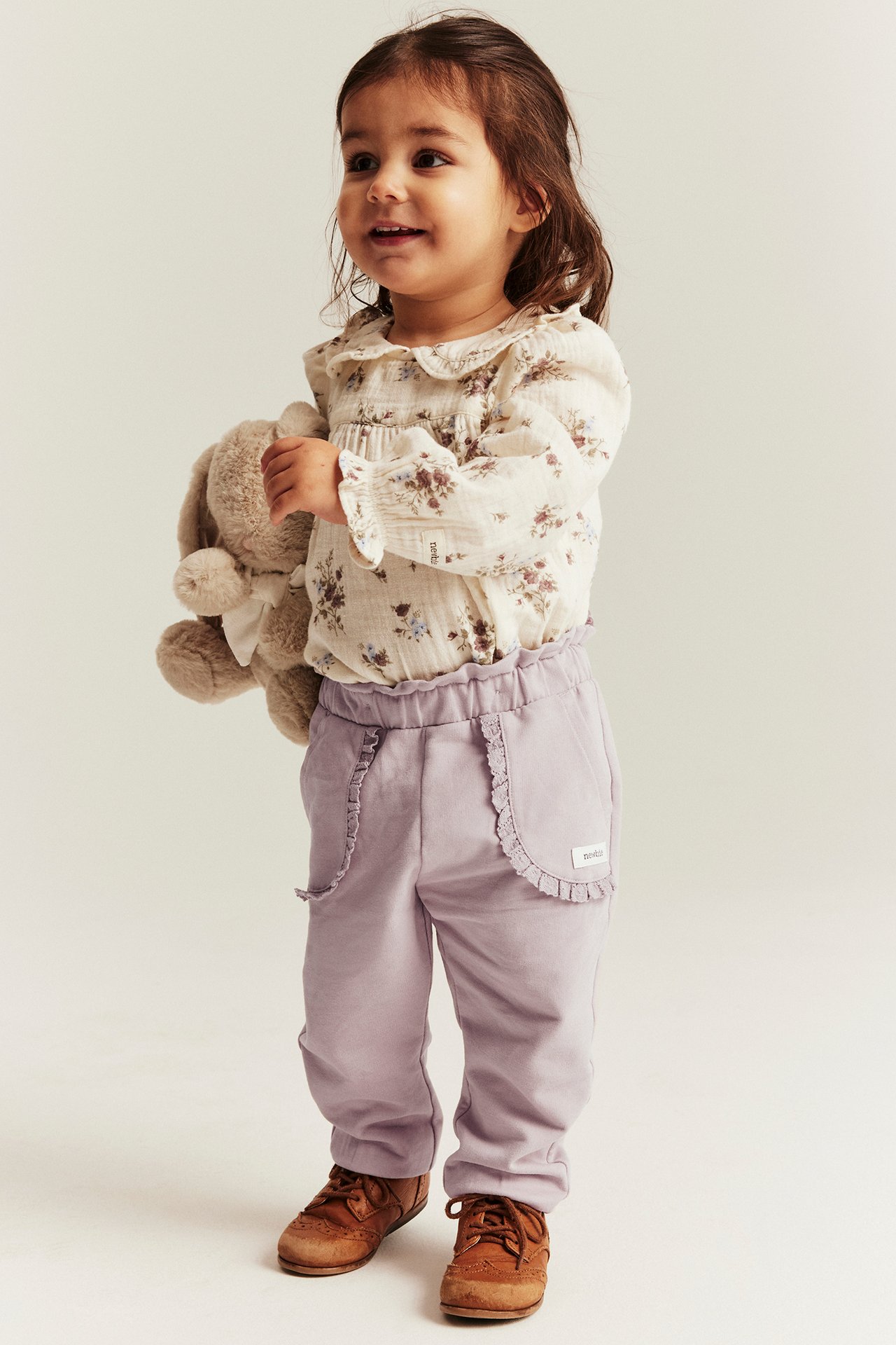 Baby girl in floral blouse, light purple pants with ruffled pockets, brown shoes, holding a beige bunny.