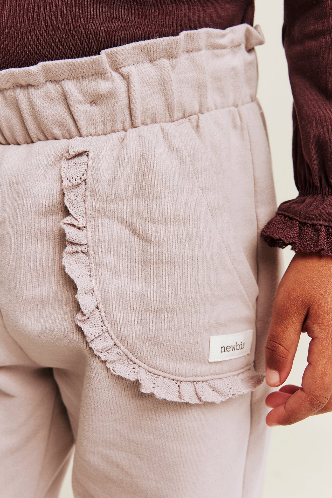 Close-up of kid wearing light purple sweatpants with a ruffled pocket and a dark brown top.