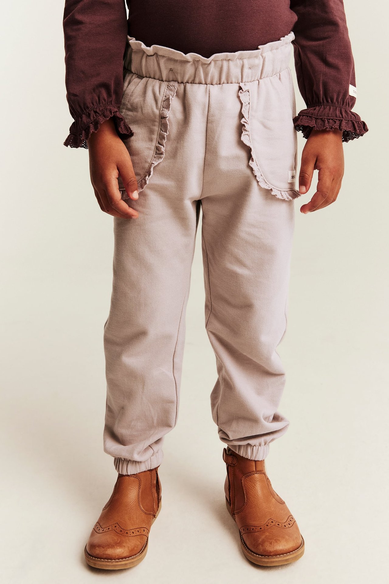 Front view of a kid wearing light brown ruffled pocket pants, a dark red ruffled top, and brown boots.