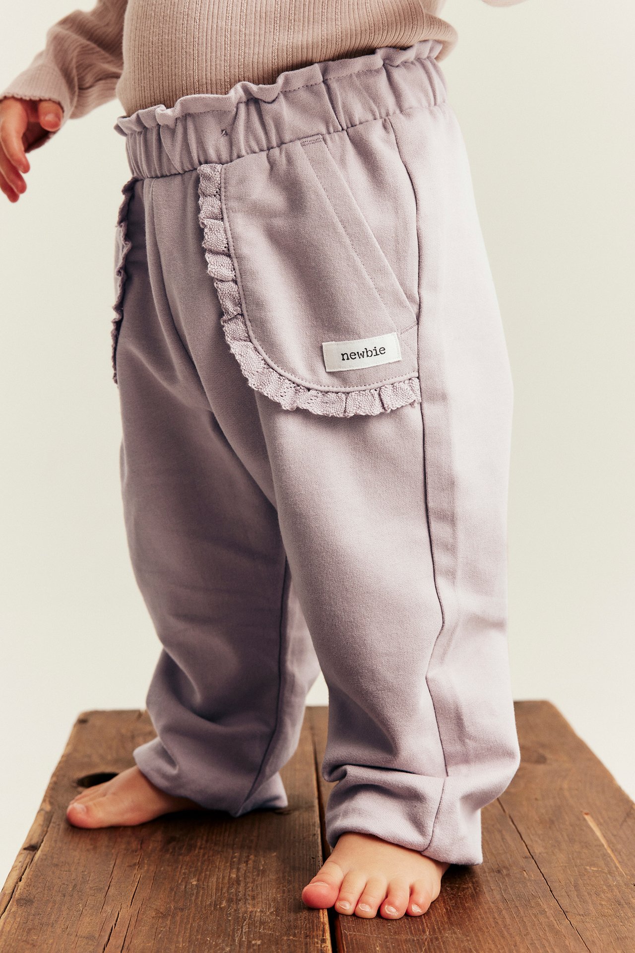 Side view of baby's light purple pants with ruffle waist and pockets, paired with a beige top.