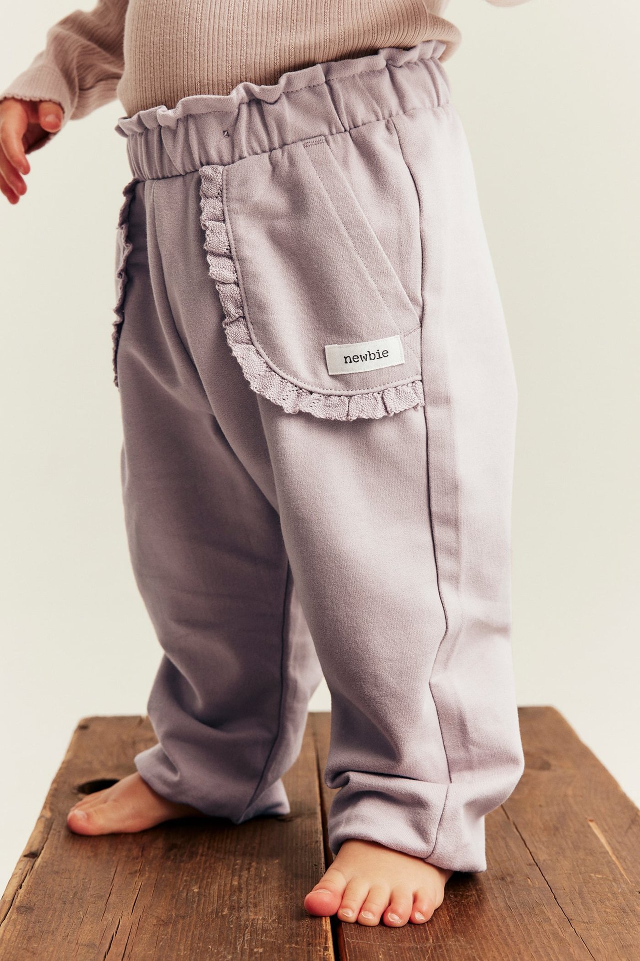 Side view of baby's light purple pants with ruffle waist and pockets, paired with a beige top.