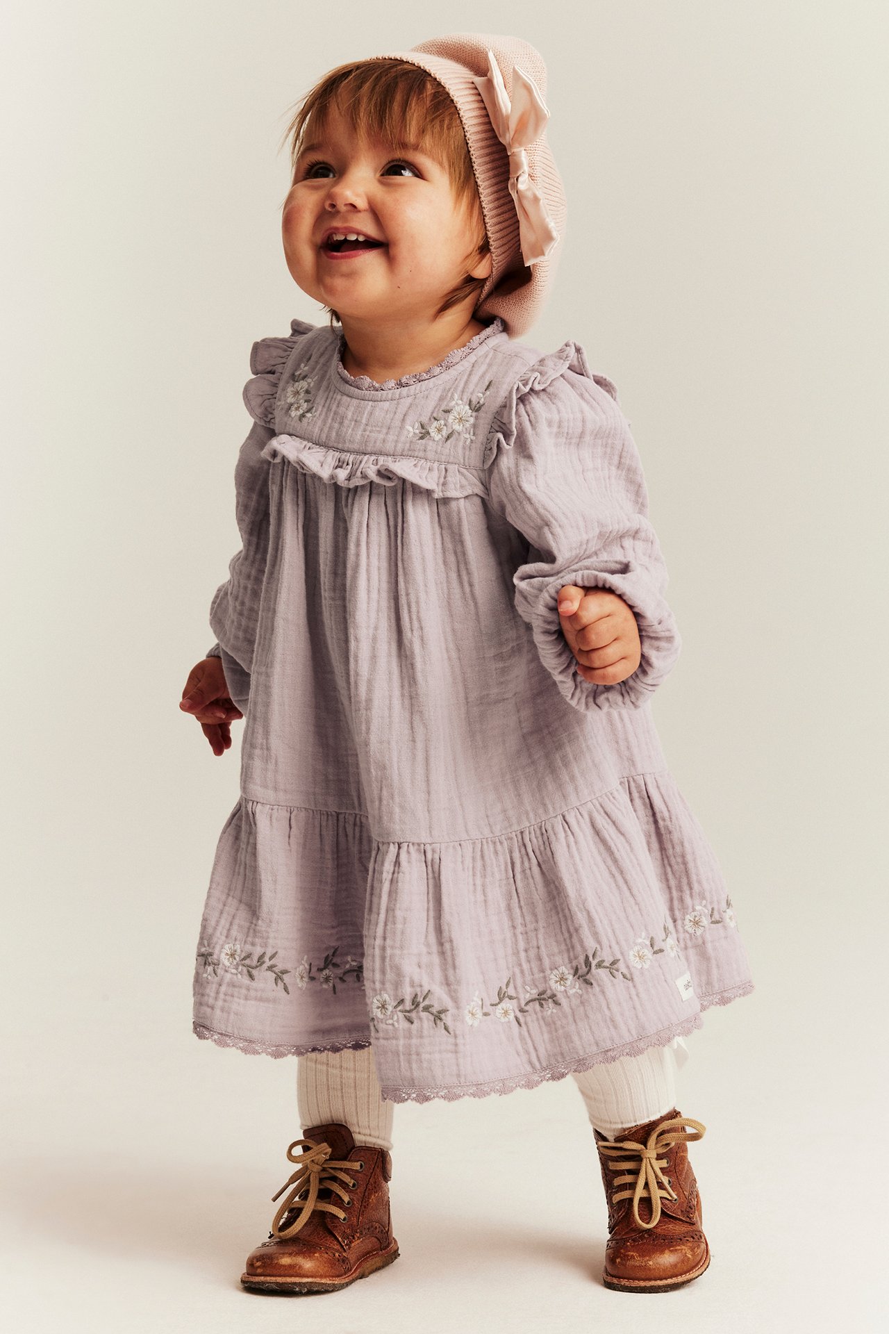 Front view: baby in a light lilac embroidered cotton dress, pink beanie, white tights, and brown boots.