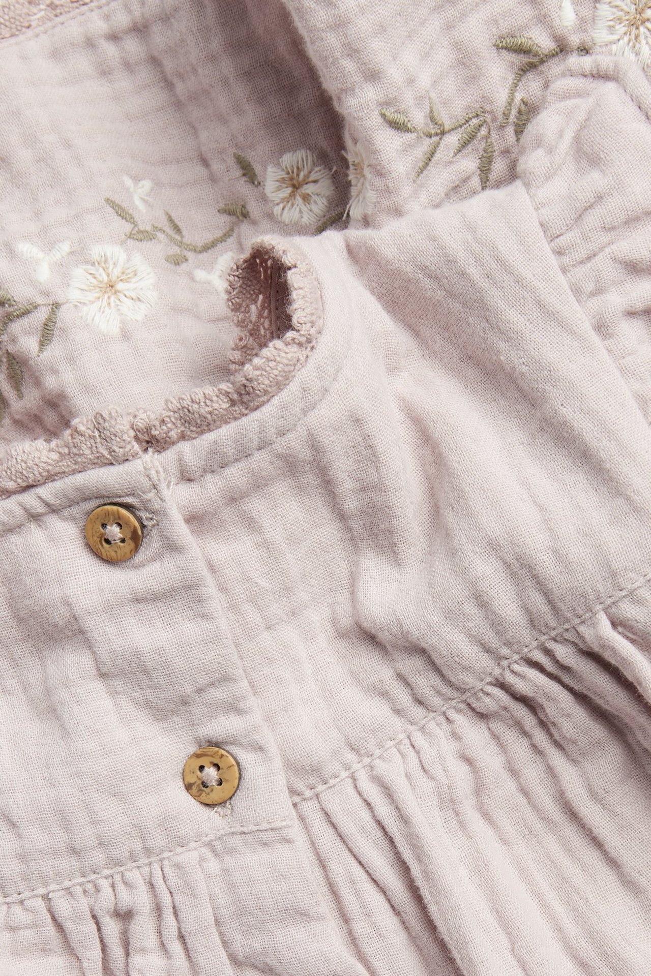 Close-up of a light lilac woven cotton dress with white floral embroidery, lace trim, and wooden buttons.