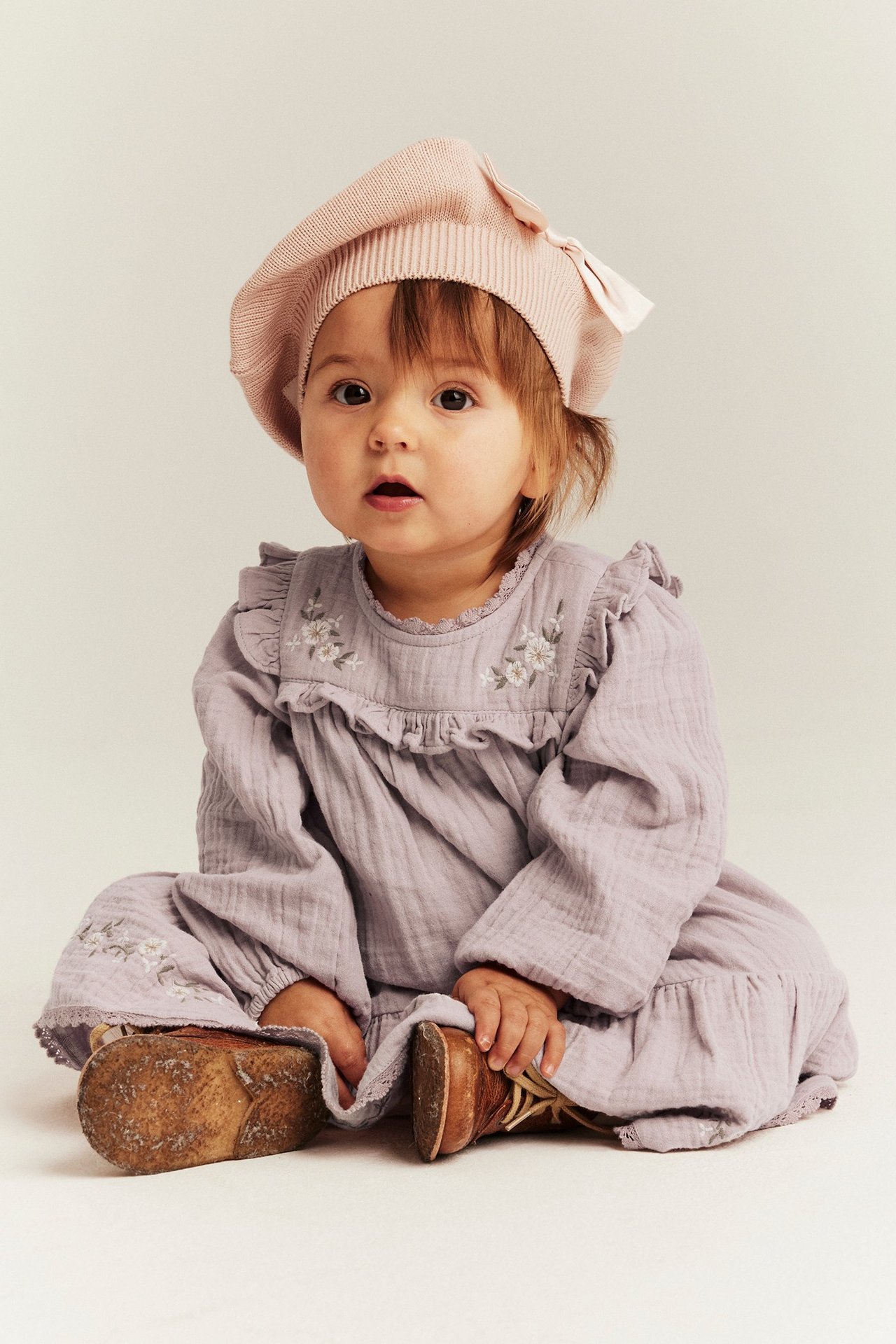 Vorderansicht: Baby trägt ein helllila gewebtes Kleid mit Blumenstickerei, Rüschen, Puffärmeln und rosa Baskenmütze.